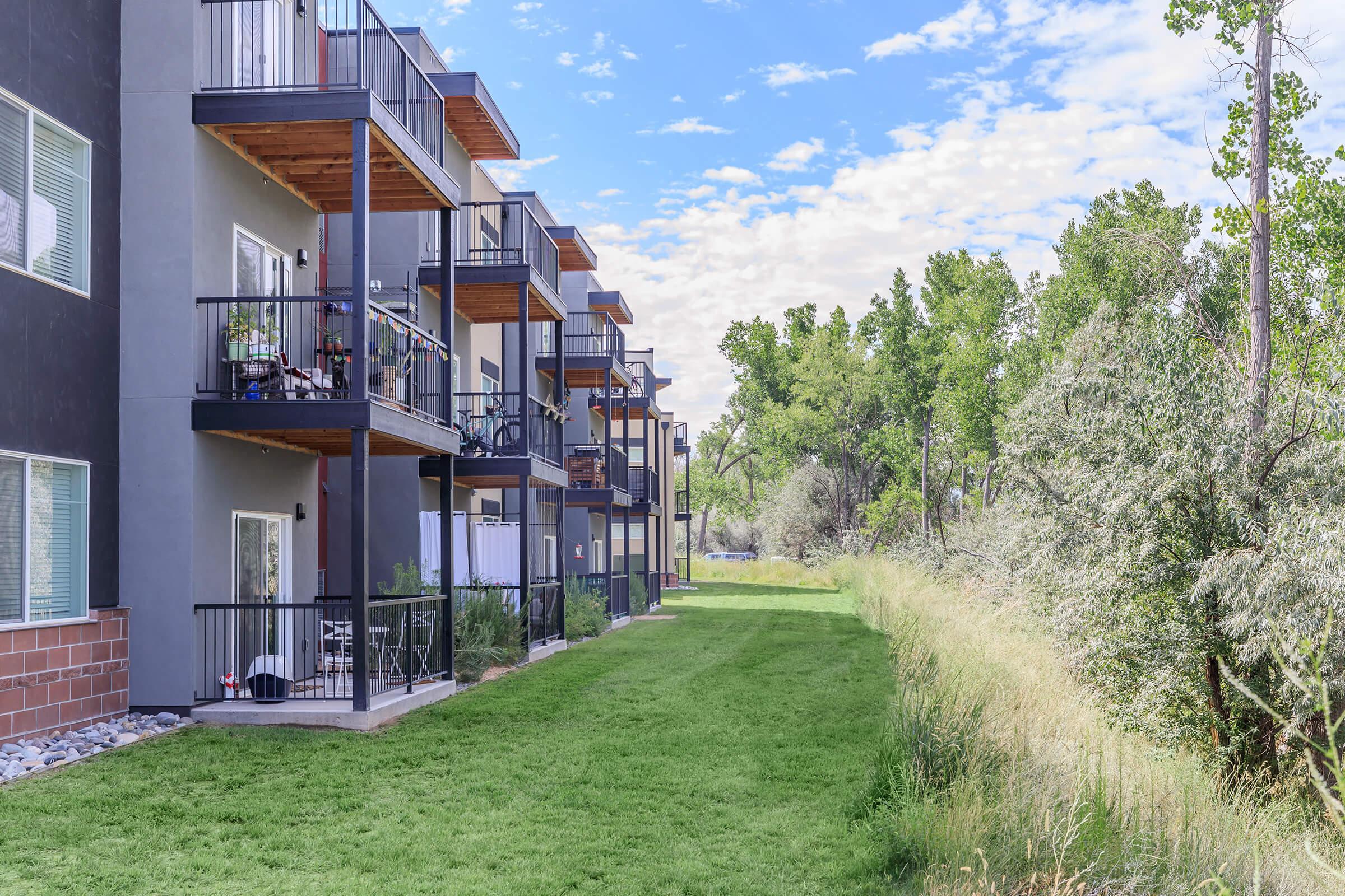 View of residential apartment buildings along a landscaped path, featuring balconies and green grass. The scene includes trees in the background and a blue sky with scattered clouds, suggesting a peaceful outdoor environment.