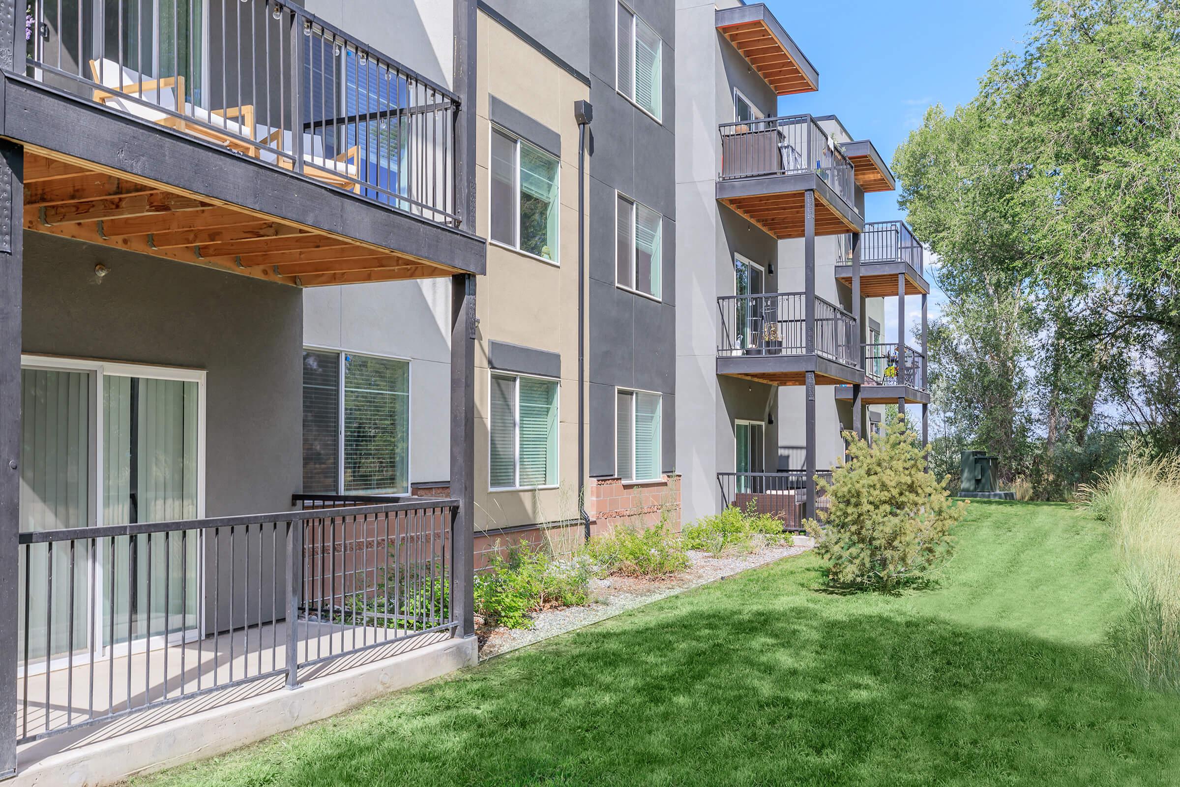 Residential apartment building featuring multiple stories, with balconies and large windows. The exterior is a mix of gray and beige, surrounded by a green lawn and small trees. The setting appears sunny and well-maintained, showcasing a peaceful living environment.
