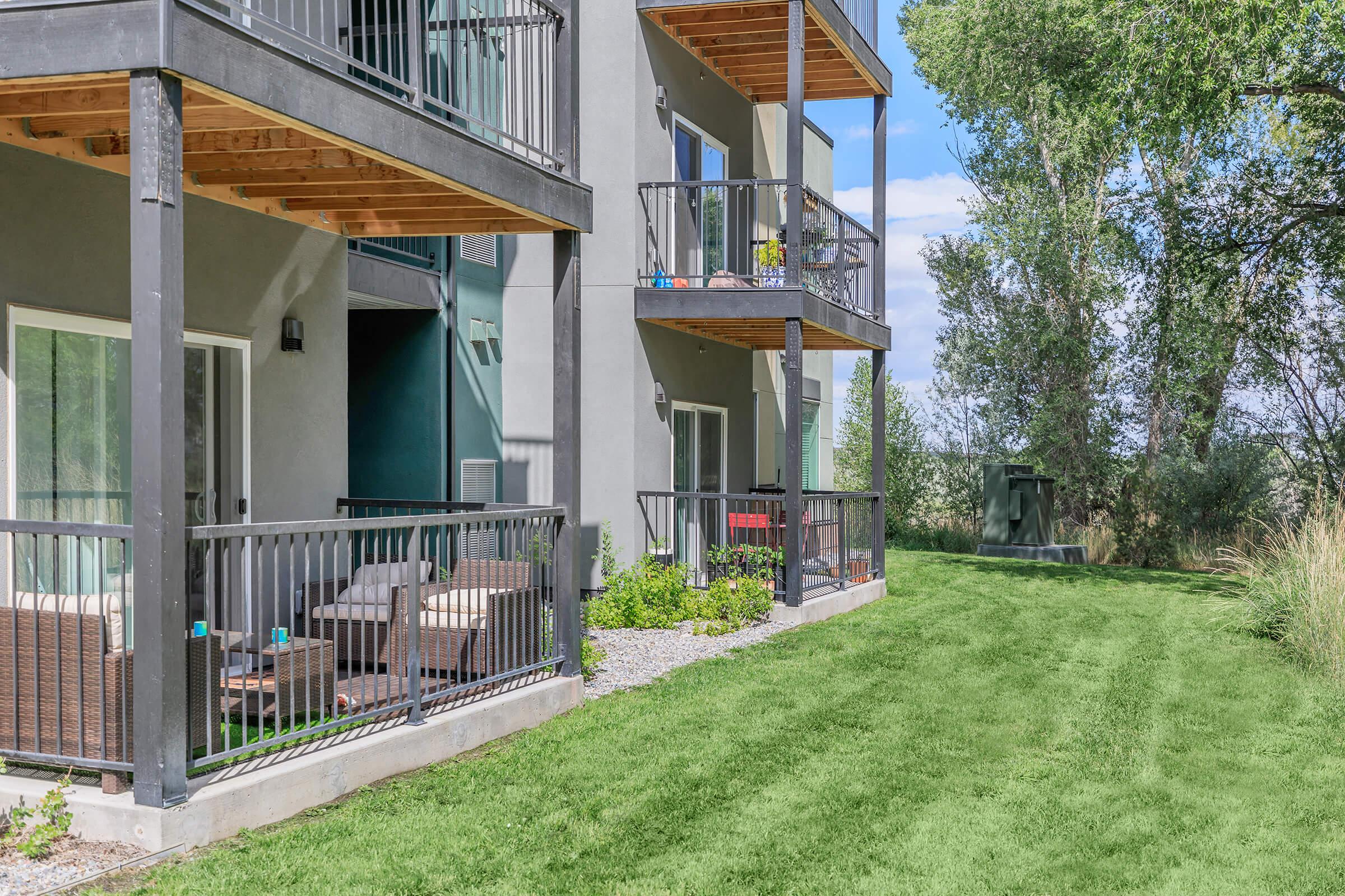 View of modern apartment balconies with wooden overhangs, surrounded by green grass and trees. Outdoor seating areas are visible on the balconies, with a well-maintained landscape in the foreground. The setting is bright and airy, suggesting a pleasant residential environment.
