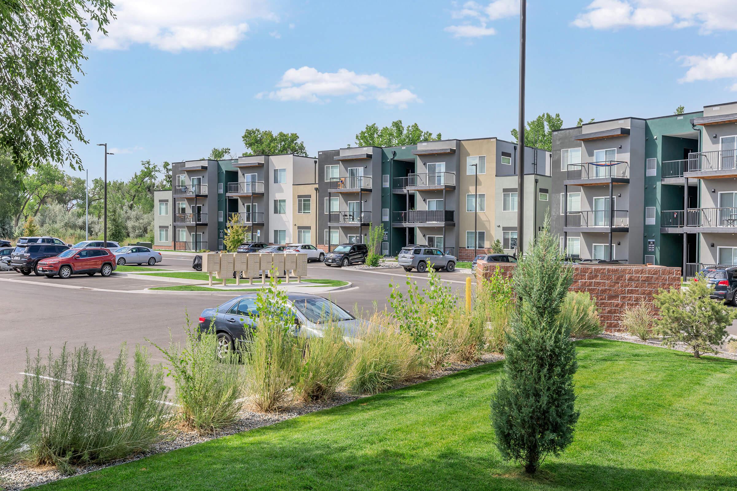A residential building complex with modern apartments is visible. The foreground features landscaped greenery, including bushes and a small tree. Multiple cars are parked in the lot, and the sky is clear with a few clouds. The overall scene conveys a suburban living environment.