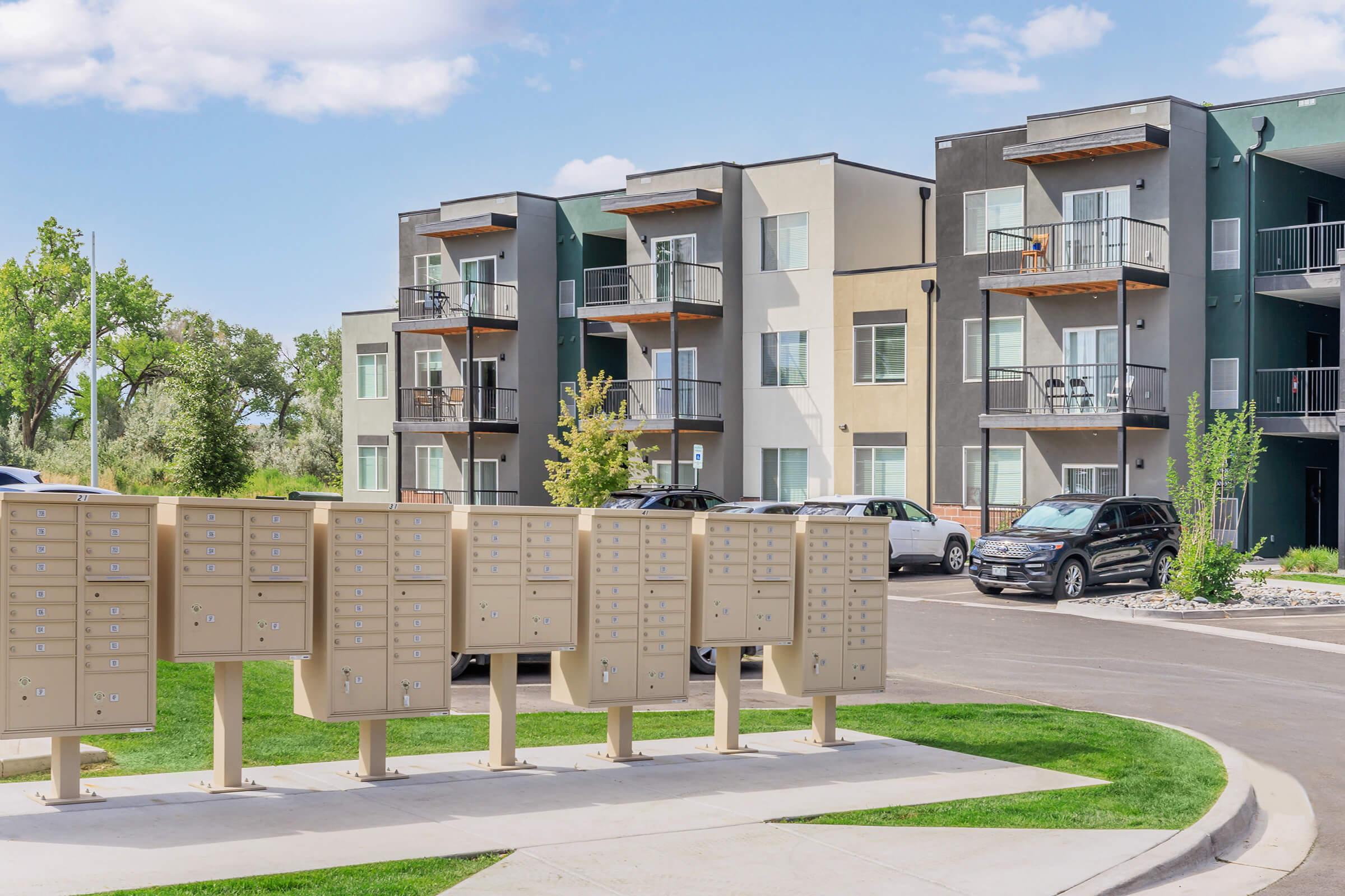 A cluster of beige mailboxes is positioned in the foreground, with modern apartment buildings in the background. The buildings feature balconies and a mix of light and dark exteriors, surrounded by greenery and parked cars. Bright blue sky with scattered clouds completes the scene.
