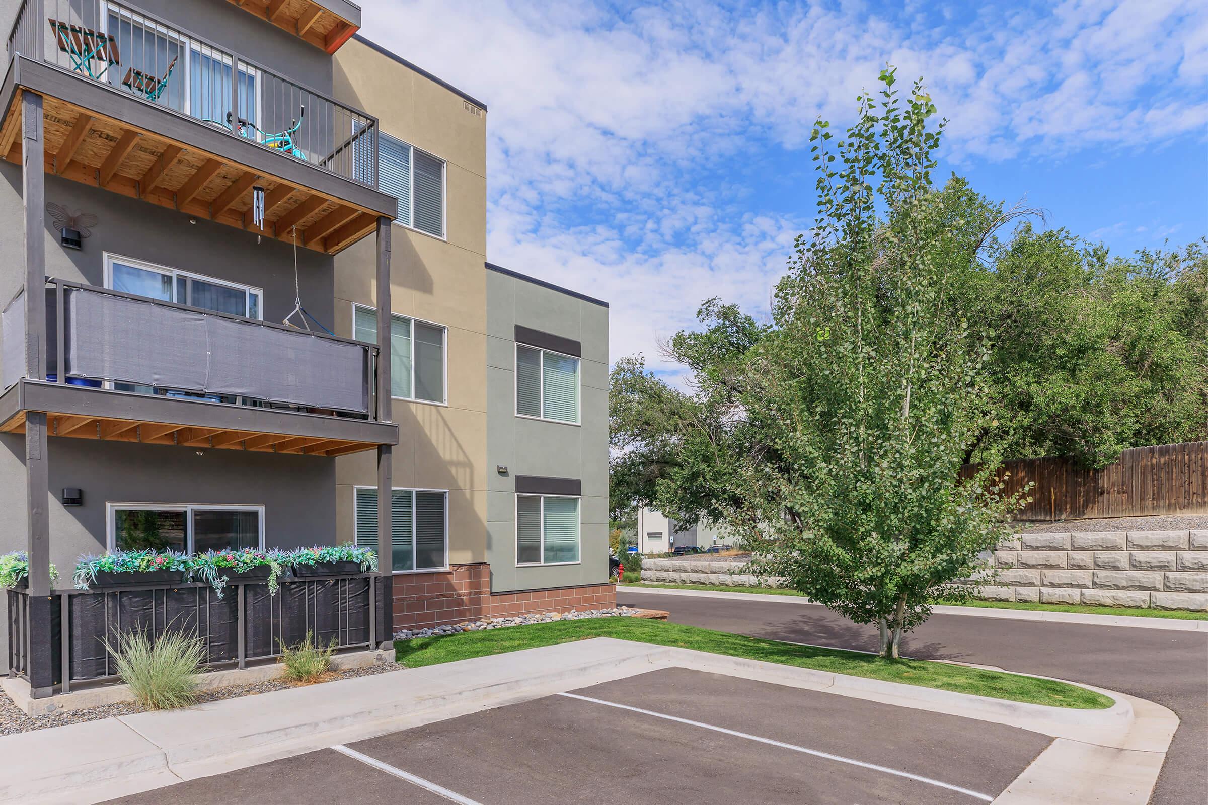 Modern apartment building with a three-story facade, featuring balconies and outdoor seating. A landscaped area with plants is visible in front. There's a clear blue sky with scattered clouds above, and a paved parking area is adjacent to the building, bordered by a neatly trimmed tree and greenery.