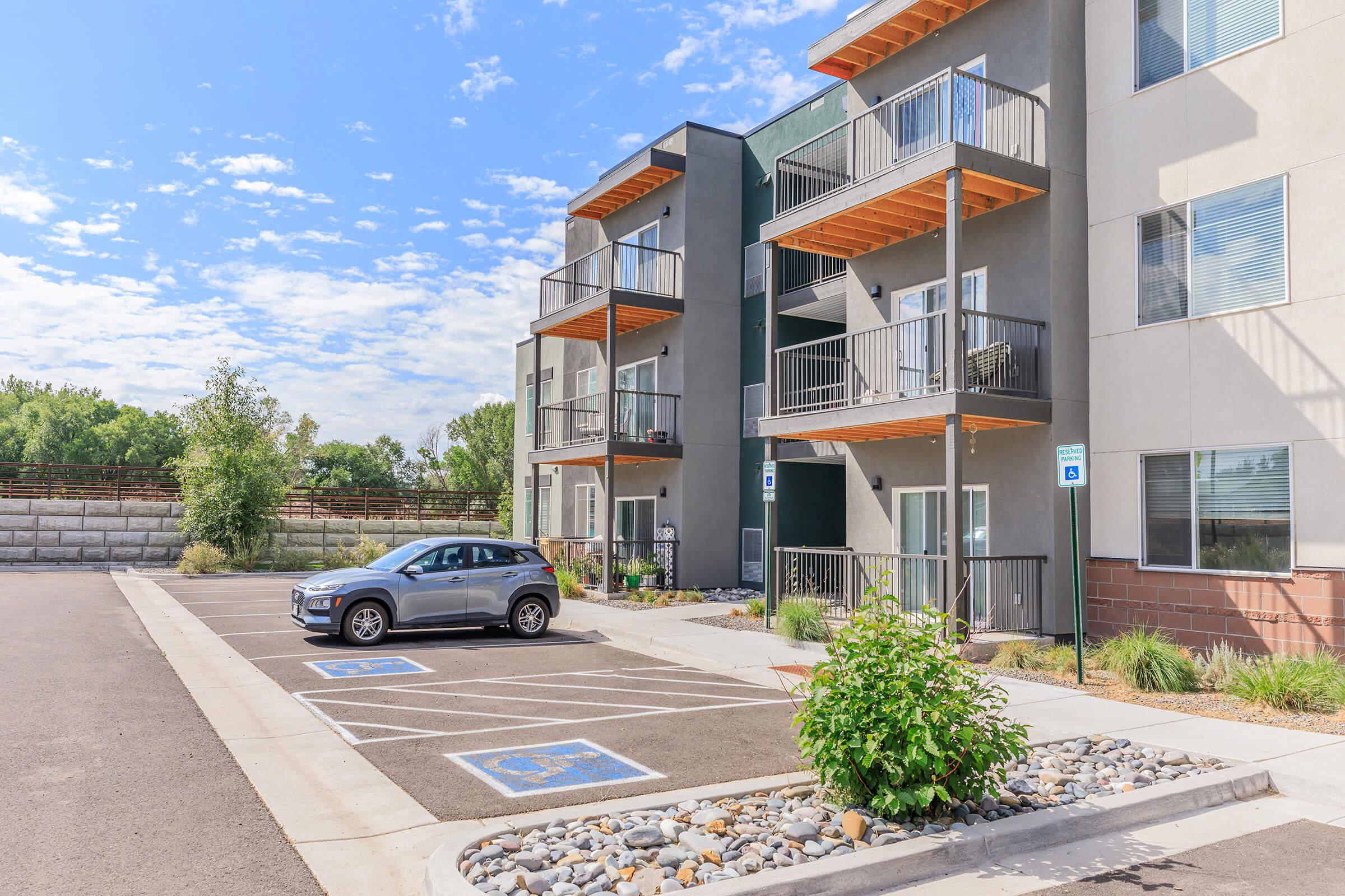 Modern apartment building with three stories, featuring balconies and large windows. A gray car is parked in a designated spot, with additional parking available. The landscape includes greenery and decorative stones, under a partly cloudy sky.