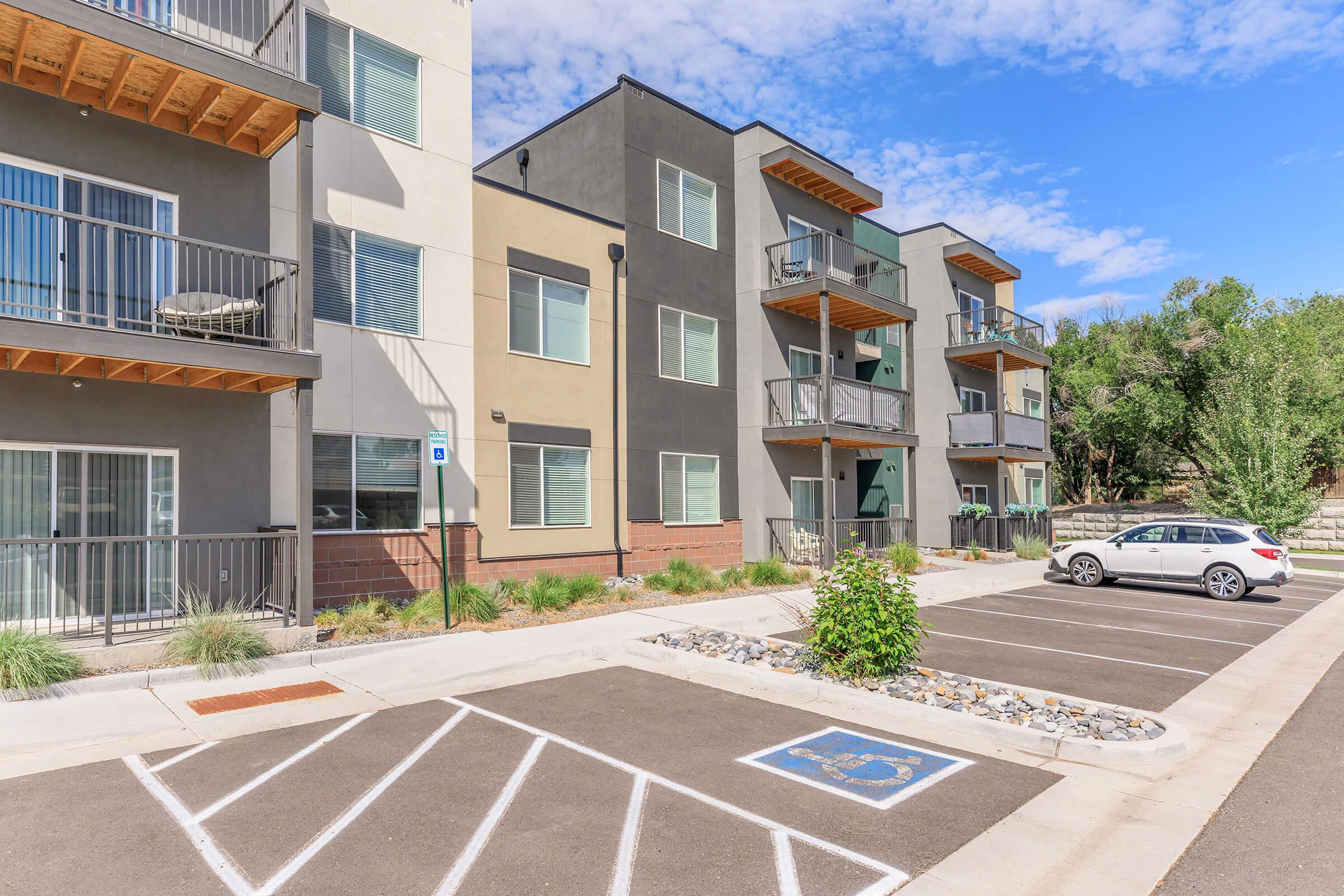 A modern apartment building with multiple floors featuring balconies and large windows. The foreground shows a parking area with marked spaces, including one for disabled parking. There are trees and shrubs along the walkway, and blue sky with scattered clouds can be seen in the background.