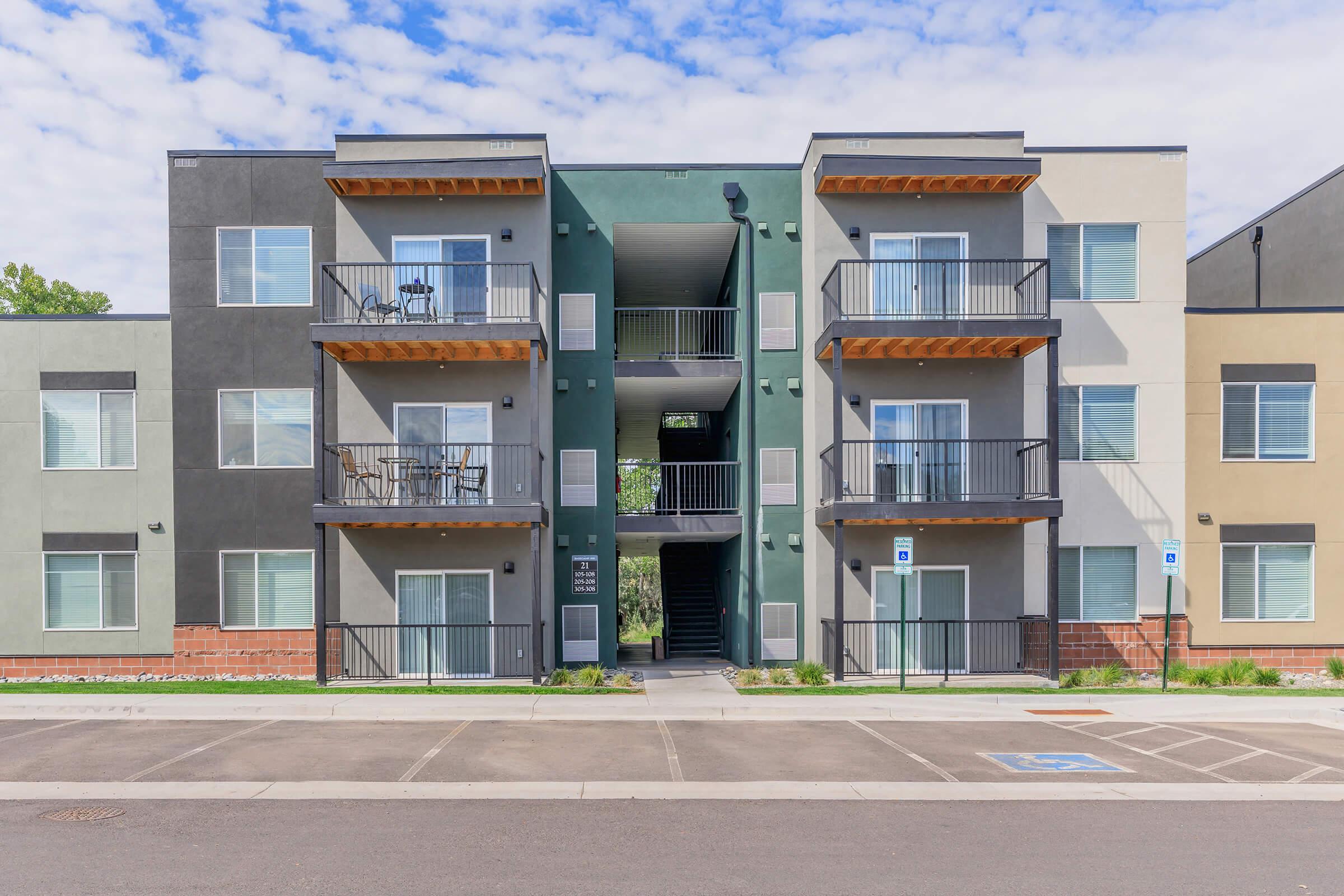 A modern three-story apartment building featuring a mix of dark and light exterior colors. Balconies with railings are visible on the upper floors. The entrance is centrally located, with an adjacent parking lot that includes designated accessible spaces. The sky is partly cloudy.
