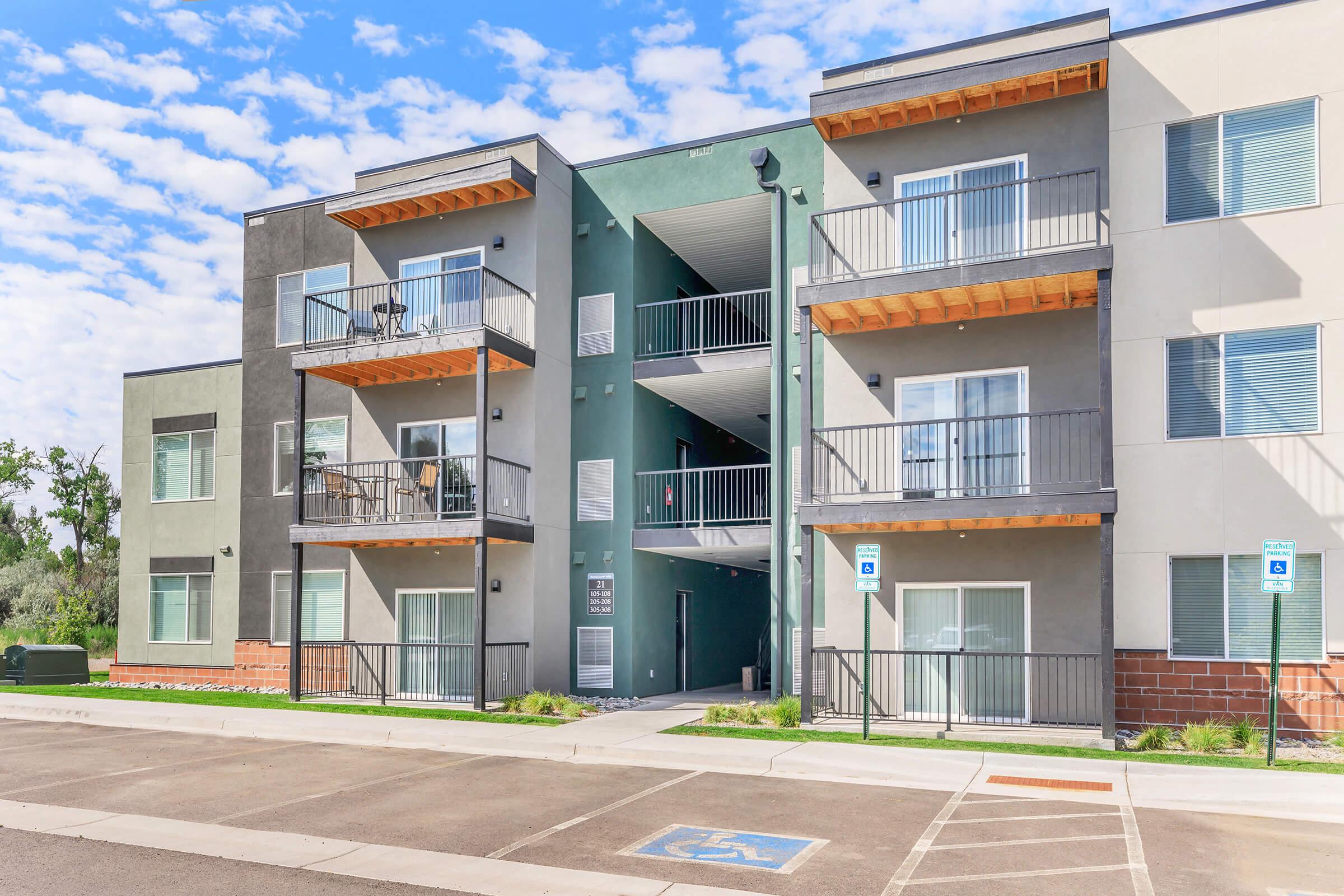 Modern apartment building with three levels, featuring balconies and a mix of green and gray exterior. The entrance has steps leading to the upper floors, and there are designated handicap parking spaces in front. The sky is partly cloudy, showcasing a bright and inviting atmosphere.