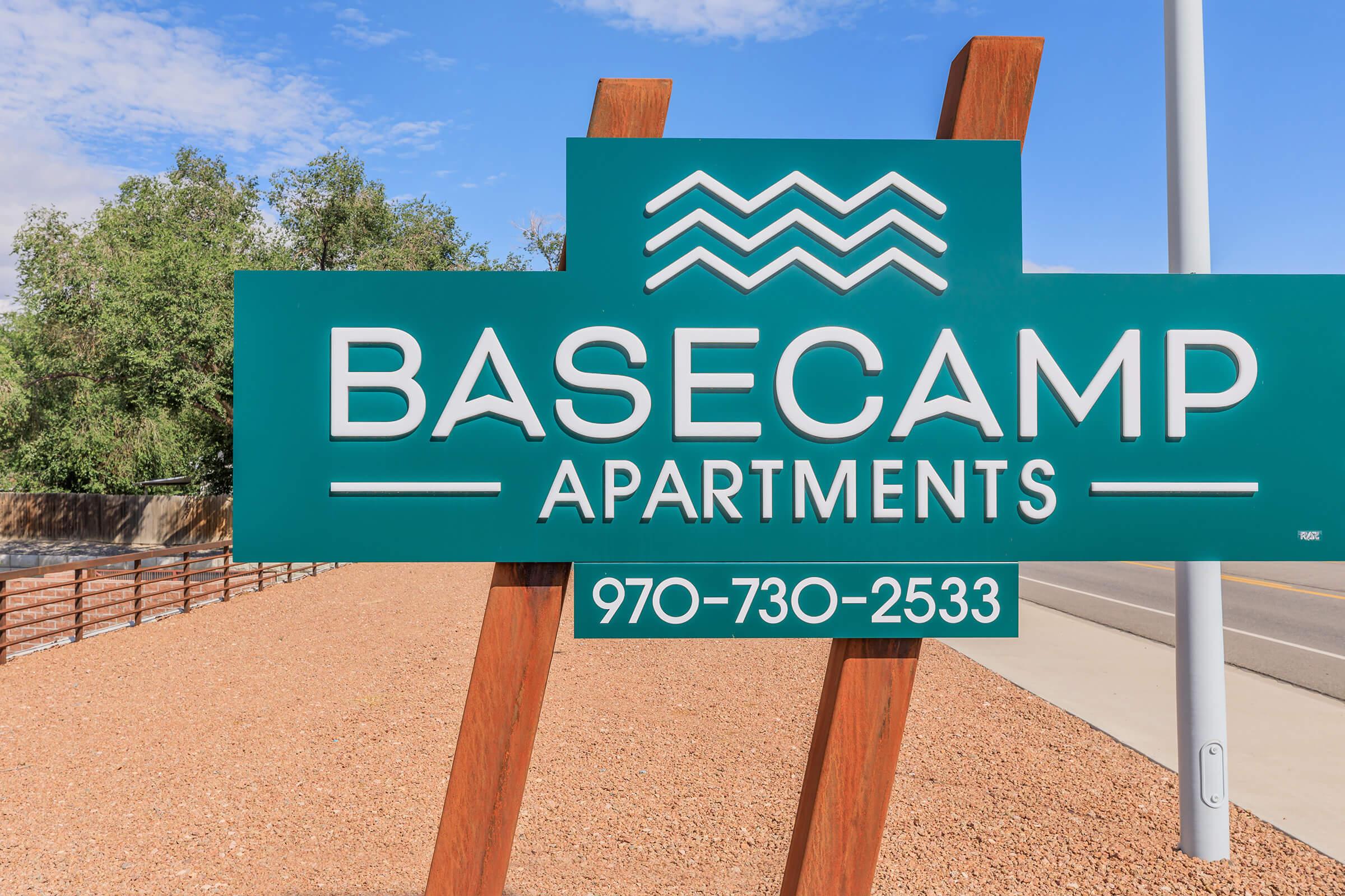 Sign for Basecamp Apartments featuring bold white lettering on a dark green background, with a design that includes wavy lines above the text. Contact number 970-730-2533 is included. The sign is positioned near a road with a gravel landscape and trees in the background under a clear blue sky.