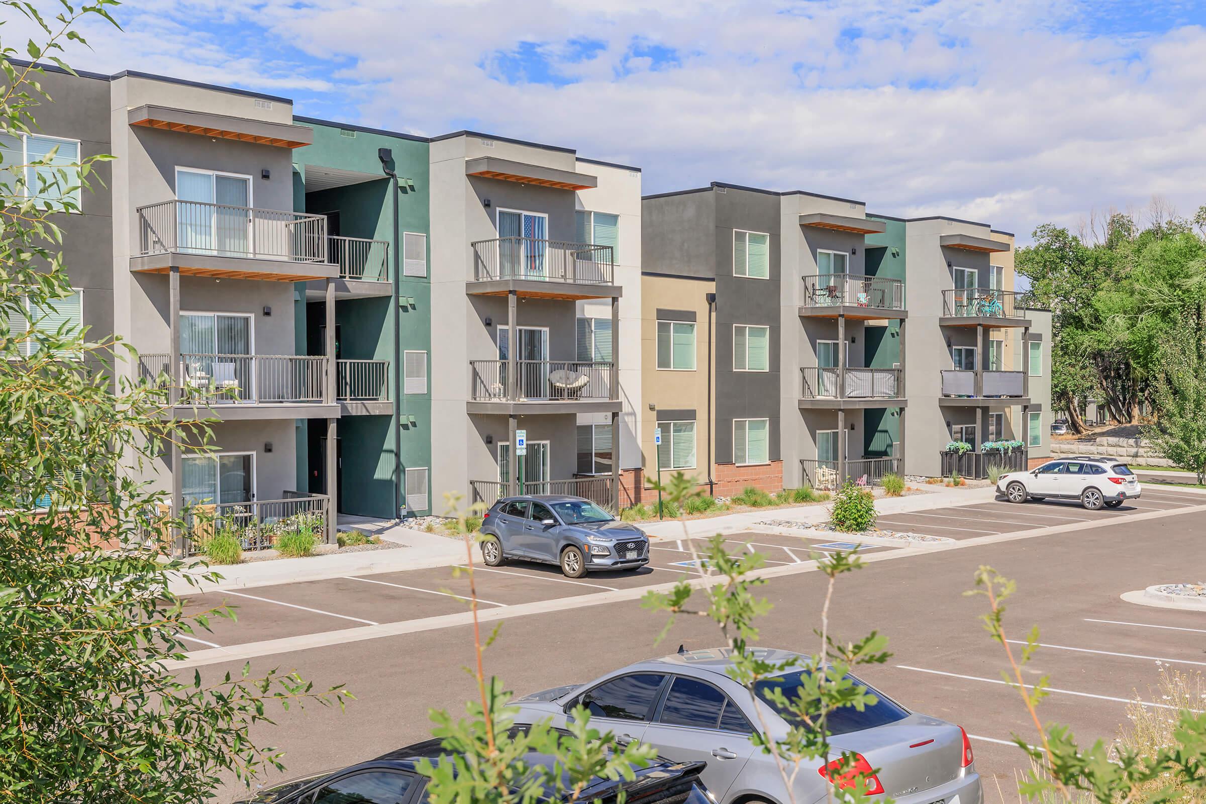A modern apartment complex featuring three-story buildings with balconies, surrounded by paved parking spaces. Several cars are parked in the lot, and greenery frames the scene against a partly cloudy sky.