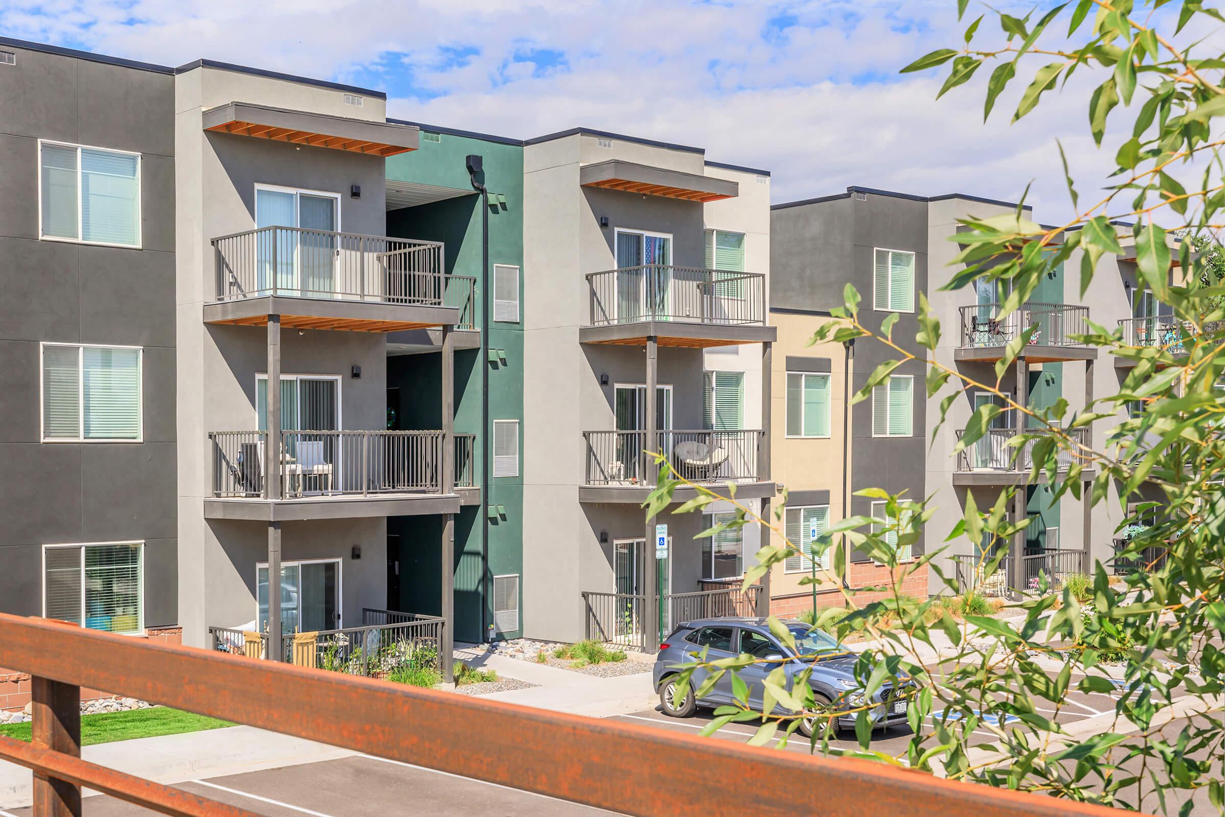 Modern apartment buildings featuring multiple balconies, with a mix of gray and beige exteriors. A tree branch is visible in the foreground, and a parked car is seen in the driveway. The scene is set under a bright blue sky with some clouds.