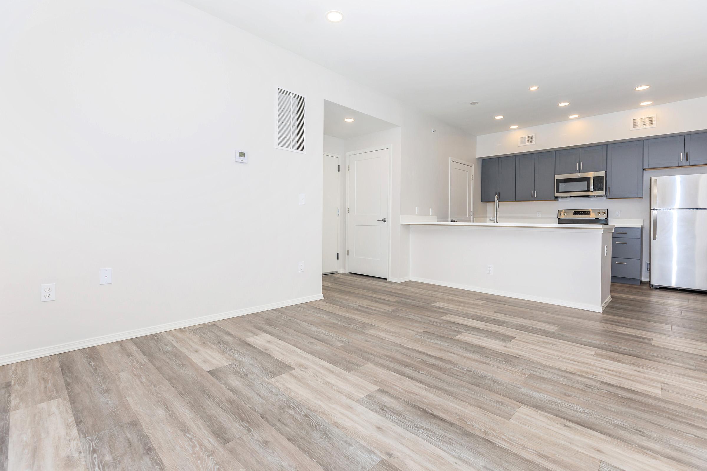 A modern, open-concept kitchen and living area featuring light-colored walls, wood-like flooring, gray cabinetry, stainless steel appliances, and ample natural light. The space is uncluttered and ready for furnishings, showcasing a contemporary design ideal for comfortable living.