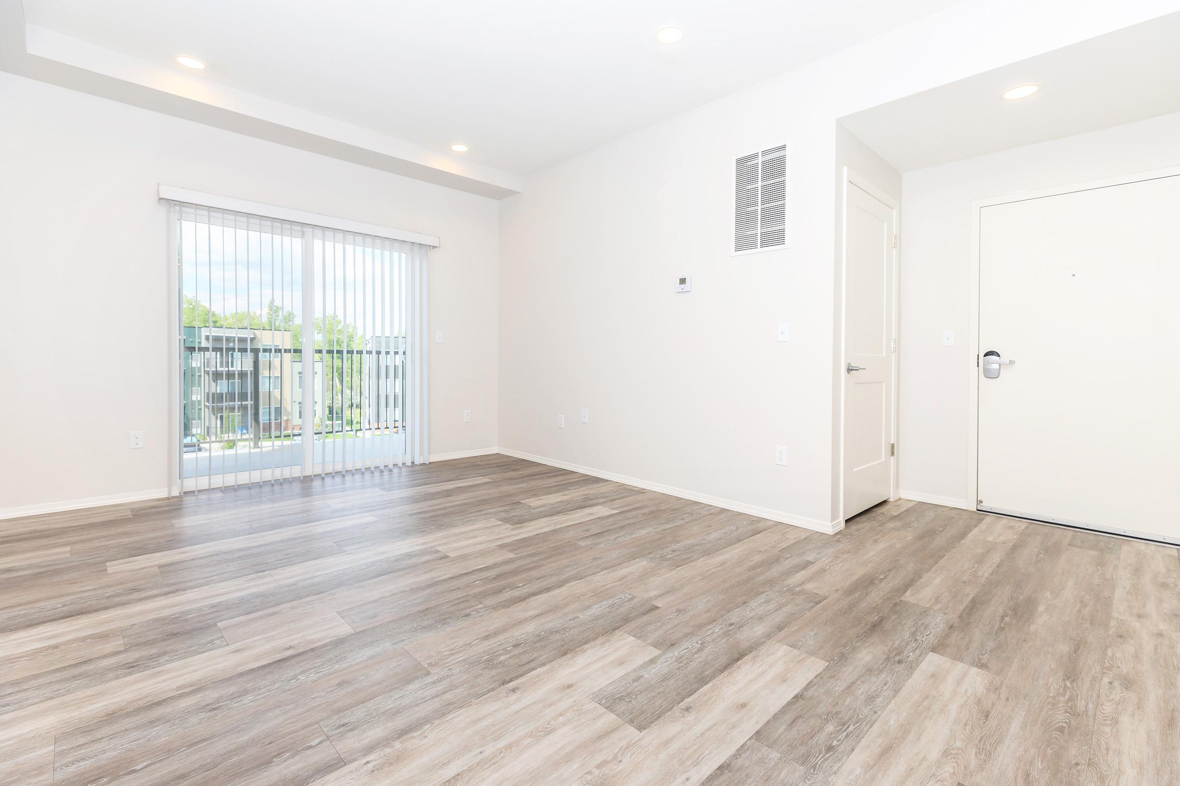 Spacious, empty living room with light-colored walls and wood-style flooring. There are large sliding glass doors with vertical blinds leading outside, and a blank wall with a small air vent. The room is well-lit with overhead lighting, creating a bright and inviting atmosphere.