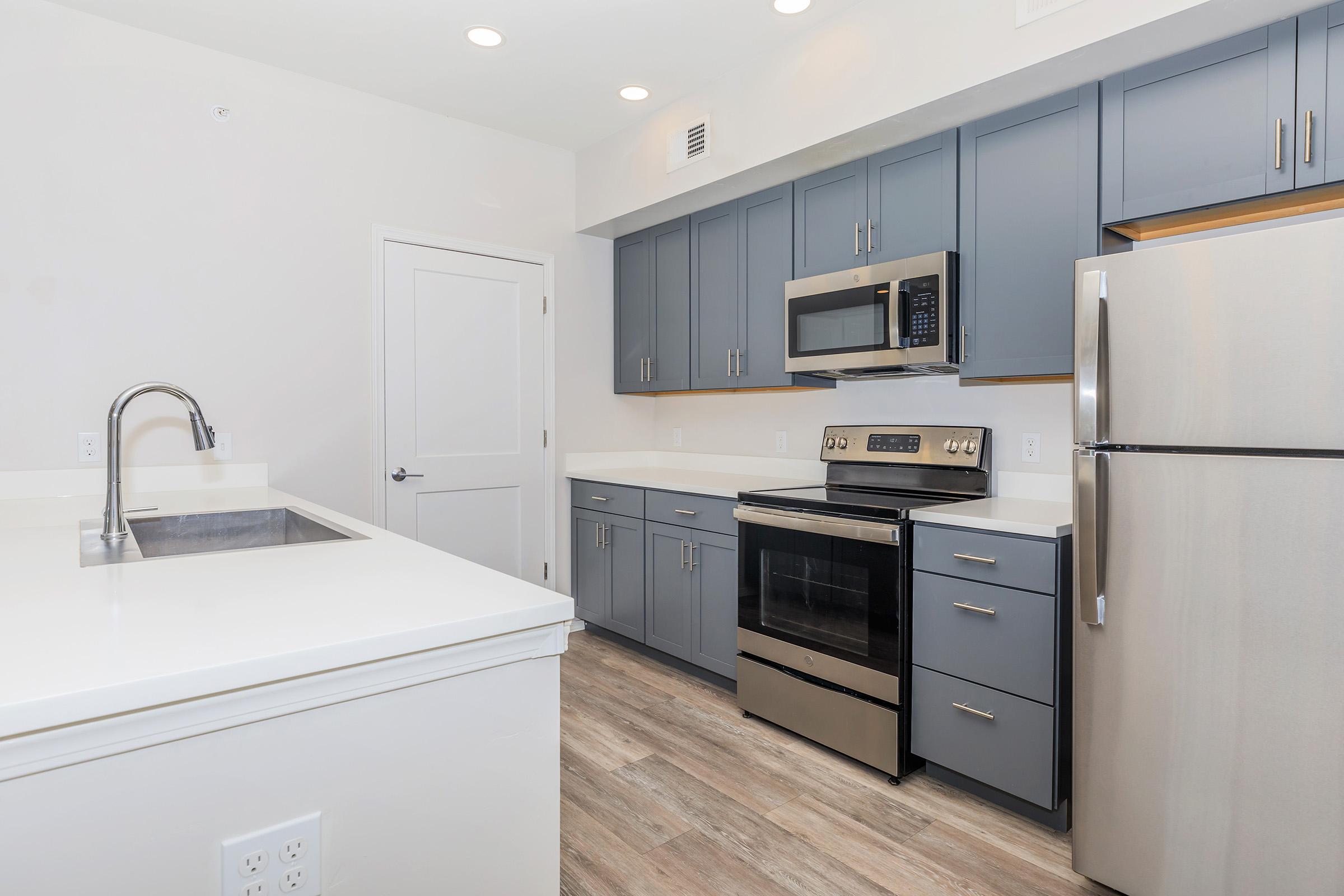 Modern kitchen featuring gray cabinets, stainless steel appliances including a microwave and a range with an oven, a silver refrigerator, a white countertop, and a sink. The space is well-lit with recessed lighting and has a light-colored wood-like floor.