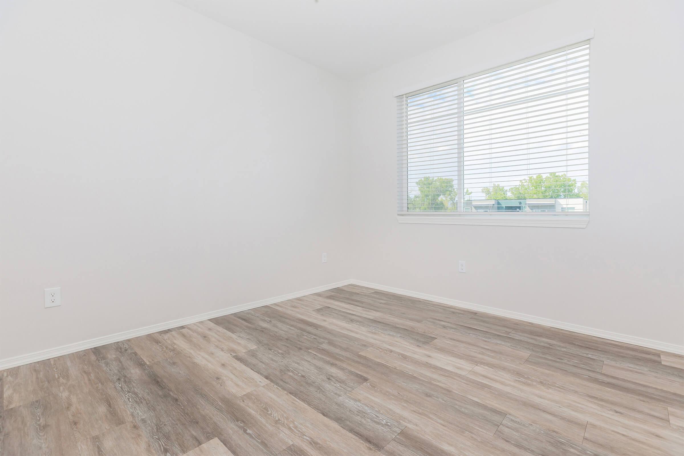 Empty room featuring light-colored walls, a large window with blinds letting in natural light, and flooring with a wood-like finish. The space is minimalistic and uncluttered, creating a bright and airy atmosphere.