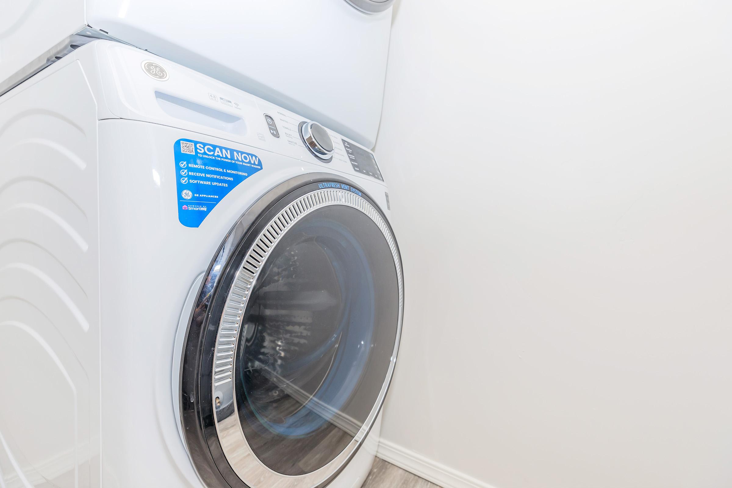 Close-up of a modern front-loading washing machine with a large circular glass door, set against a light-colored wall. A blue sticker on the machine reads "SCAN NOW." The image features a clean and minimalistic laundry space.