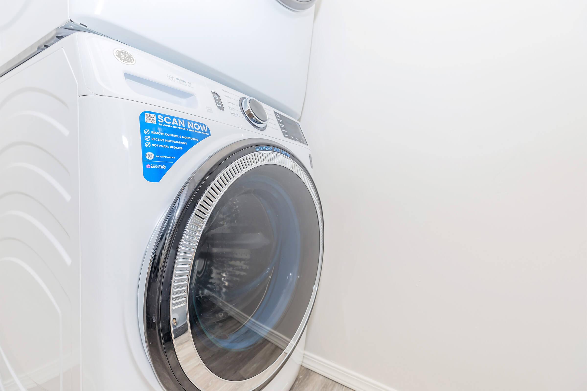 A modern front-loading washing machine in a light-colored laundry room. The machine features a blue "Scan Now" sticker on the front and has a shiny circular door showcasing its interior. The surrounding walls are plain and the floor is wooden, creating a clean and minimalist appearance.