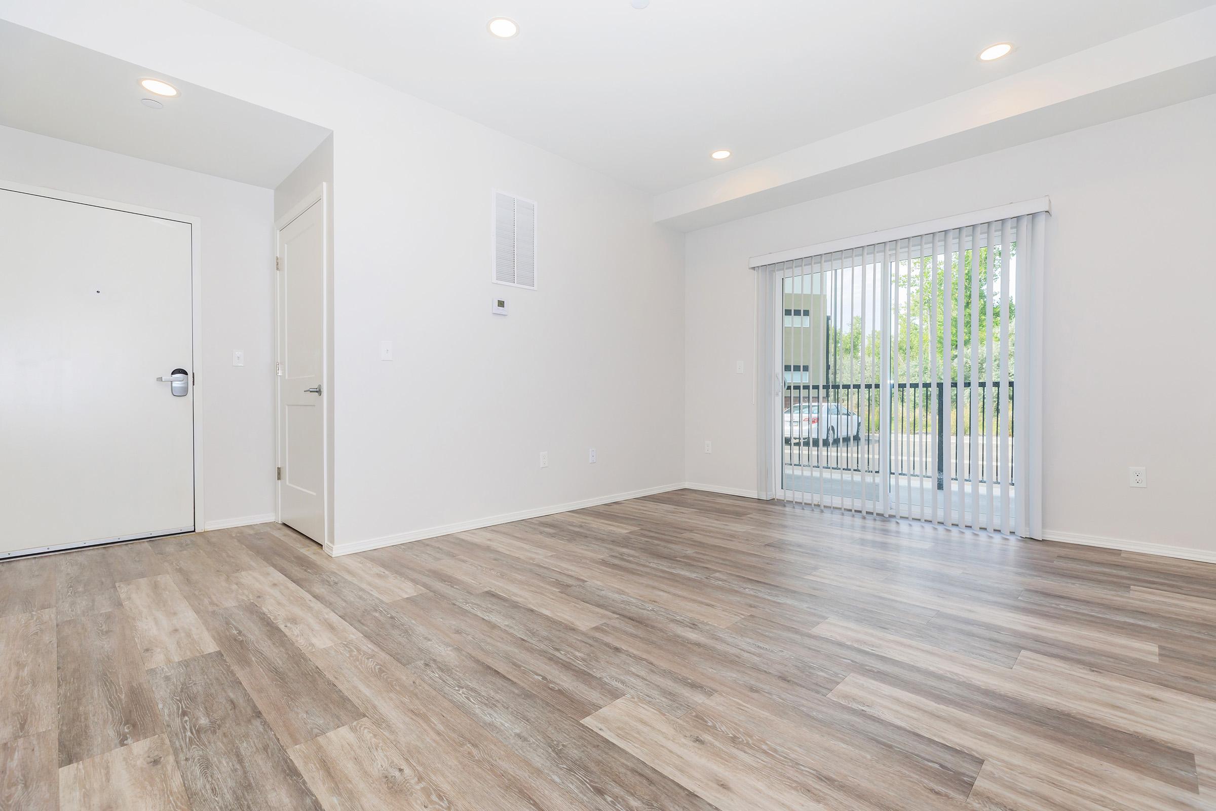 A spacious, empty room with light-colored walls and wood laminate flooring. A large window with vertical blinds allows natural light to enter, and a door leads outside. The room is well-lit, featuring recessed ceiling lights, and has a clean, modern aesthetic.
