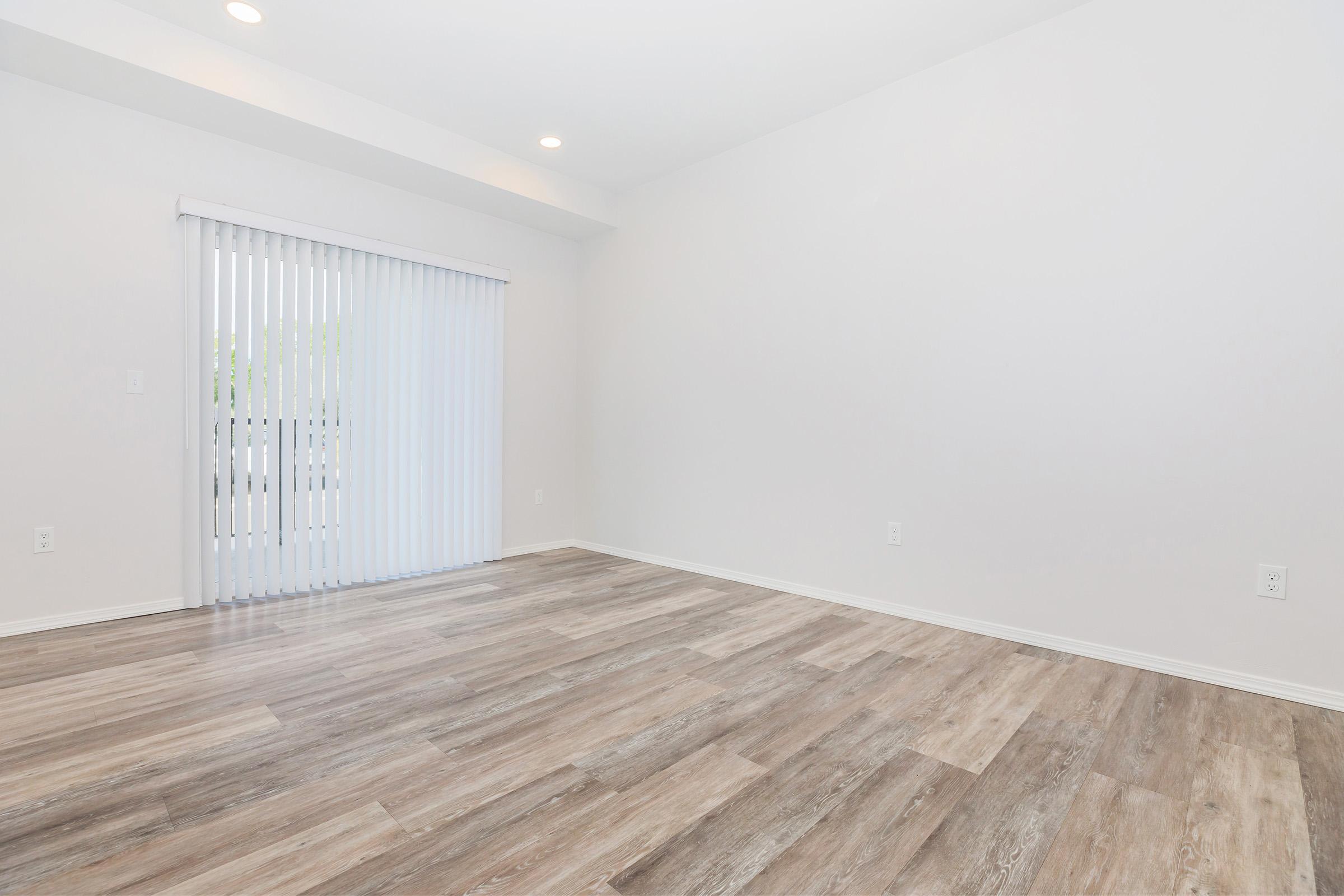 Empty room with light-colored walls and wooden flooring. A set of vertical blinds covers a window, allowing natural light to enter. The space is unfurnished, creating a clean and open atmosphere.