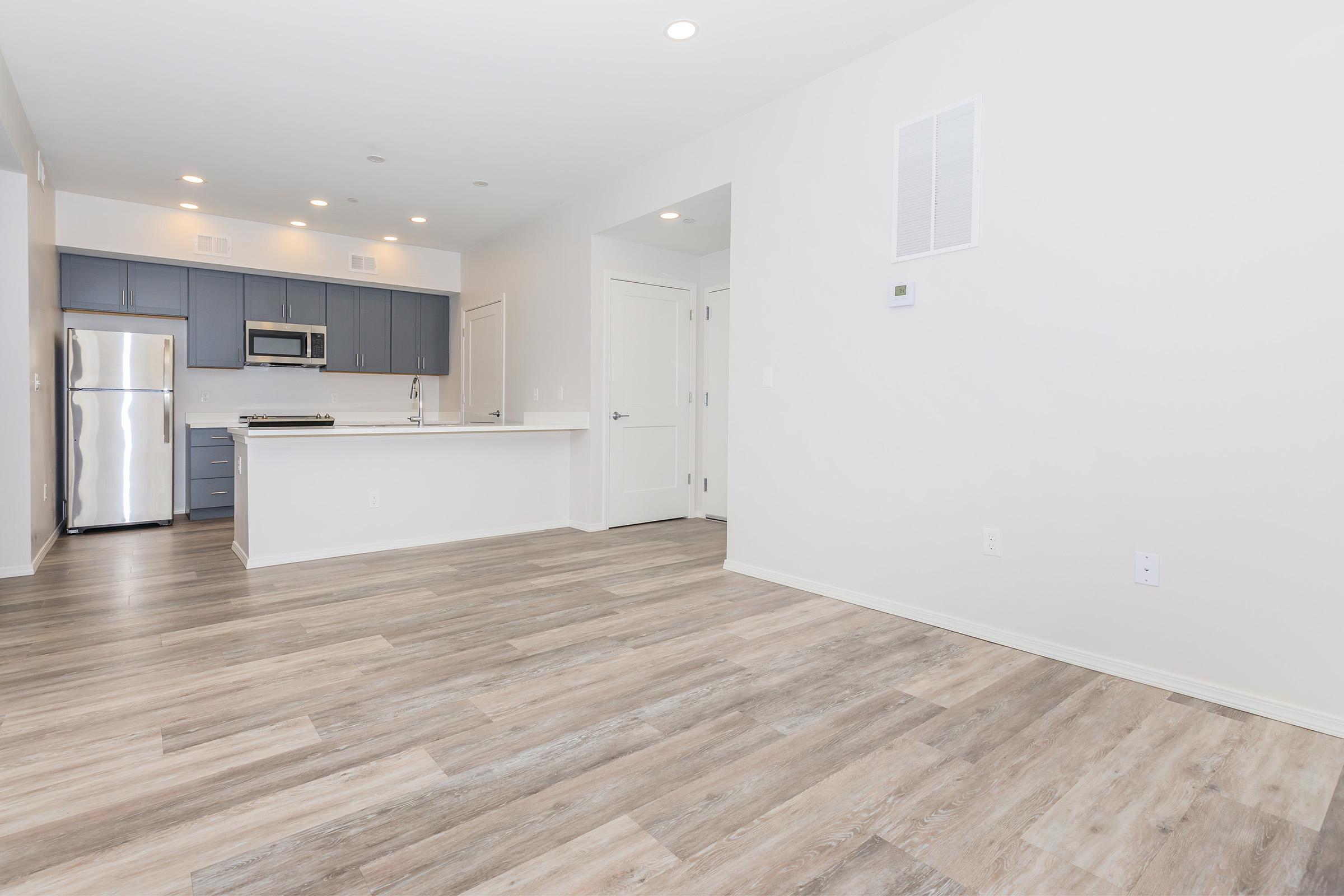Modern interior of an empty living space featuring hardwood flooring, a kitchen with stainless steel appliances, and gray cabinetry. The walls are painted white, creating a bright and open atmosphere. Natural light comes in through overhead lights and large windows.