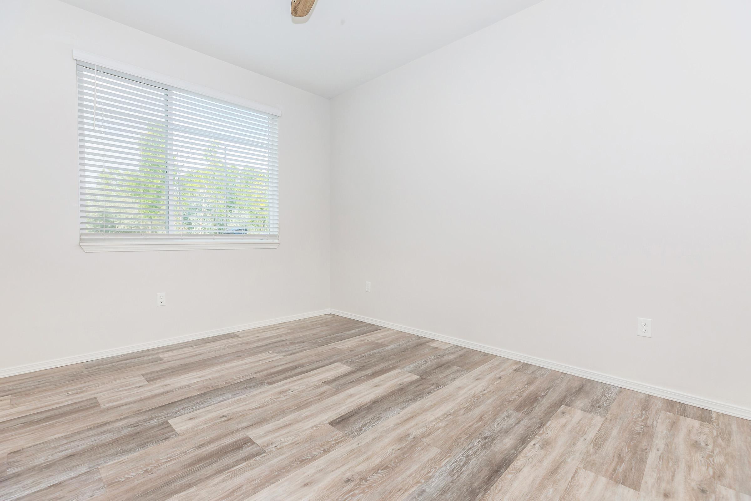 Empty room with light-colored walls and a wooden floor. A window with white blinds lets in natural light, illuminating the space. The room appears clean and minimalist, providing a blank canvas for decoration or furnishing.