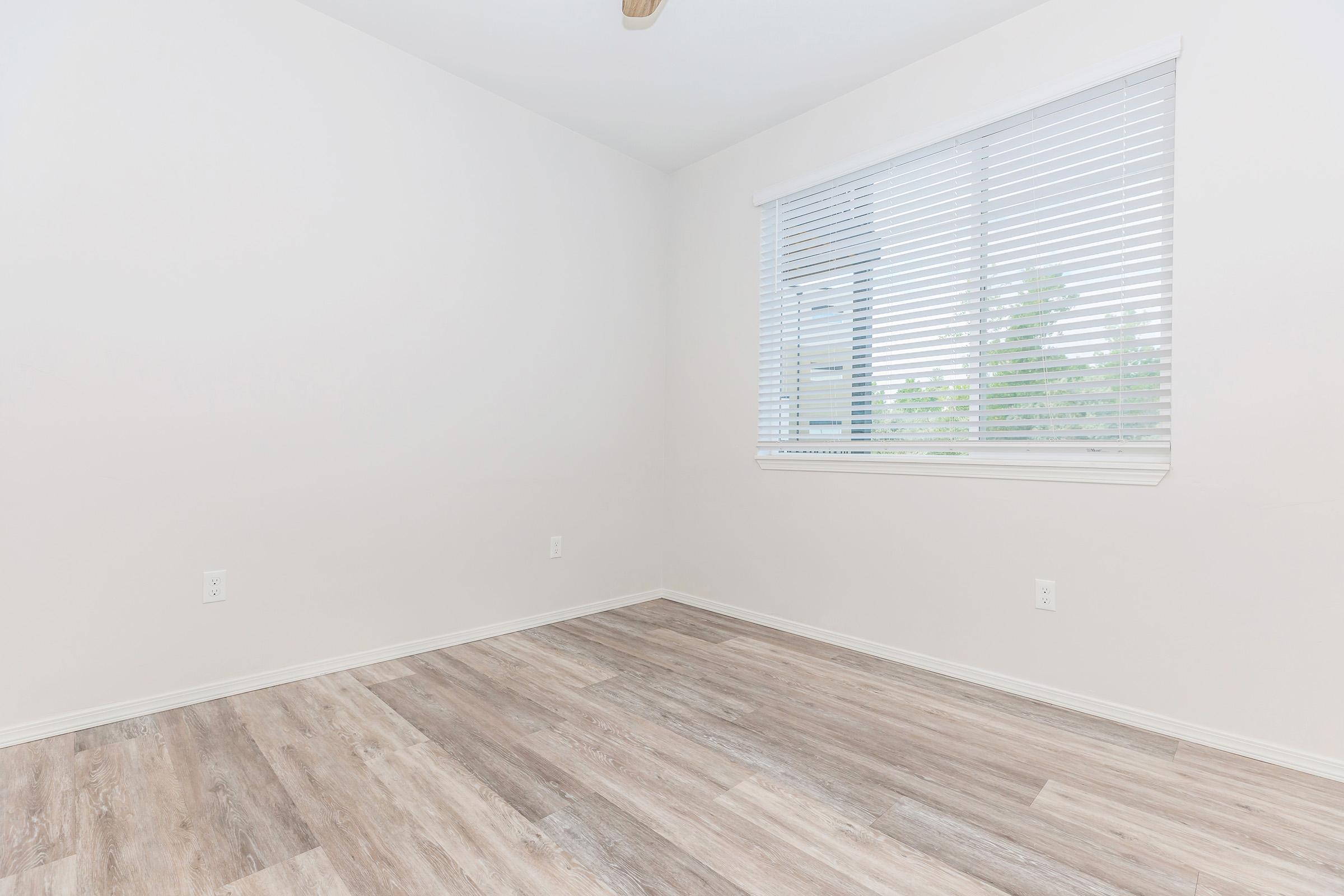 A minimalist room featuring light-colored walls and a hardwood floor. A window with blinds allows natural light to enter, illuminating the empty space. The room is unfurnished, creating a bright and open atmosphere, ideal for various uses or decorating styles.