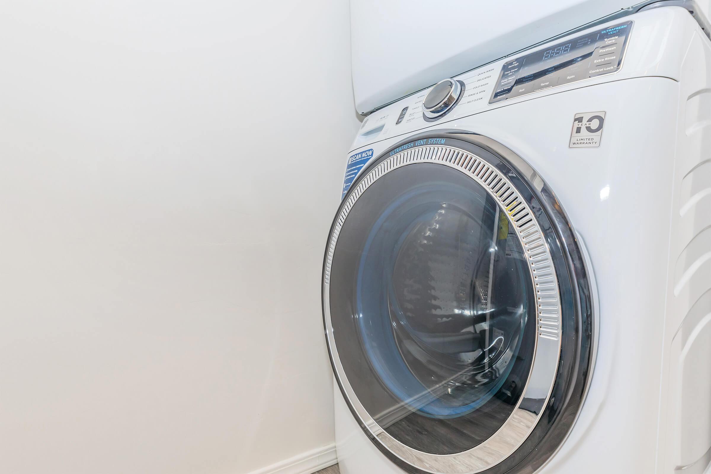 A modern front-loading washing machine with a transparent door, showing a drum and control panel. The machine is white with chrome accents, situated against a light-colored wall.