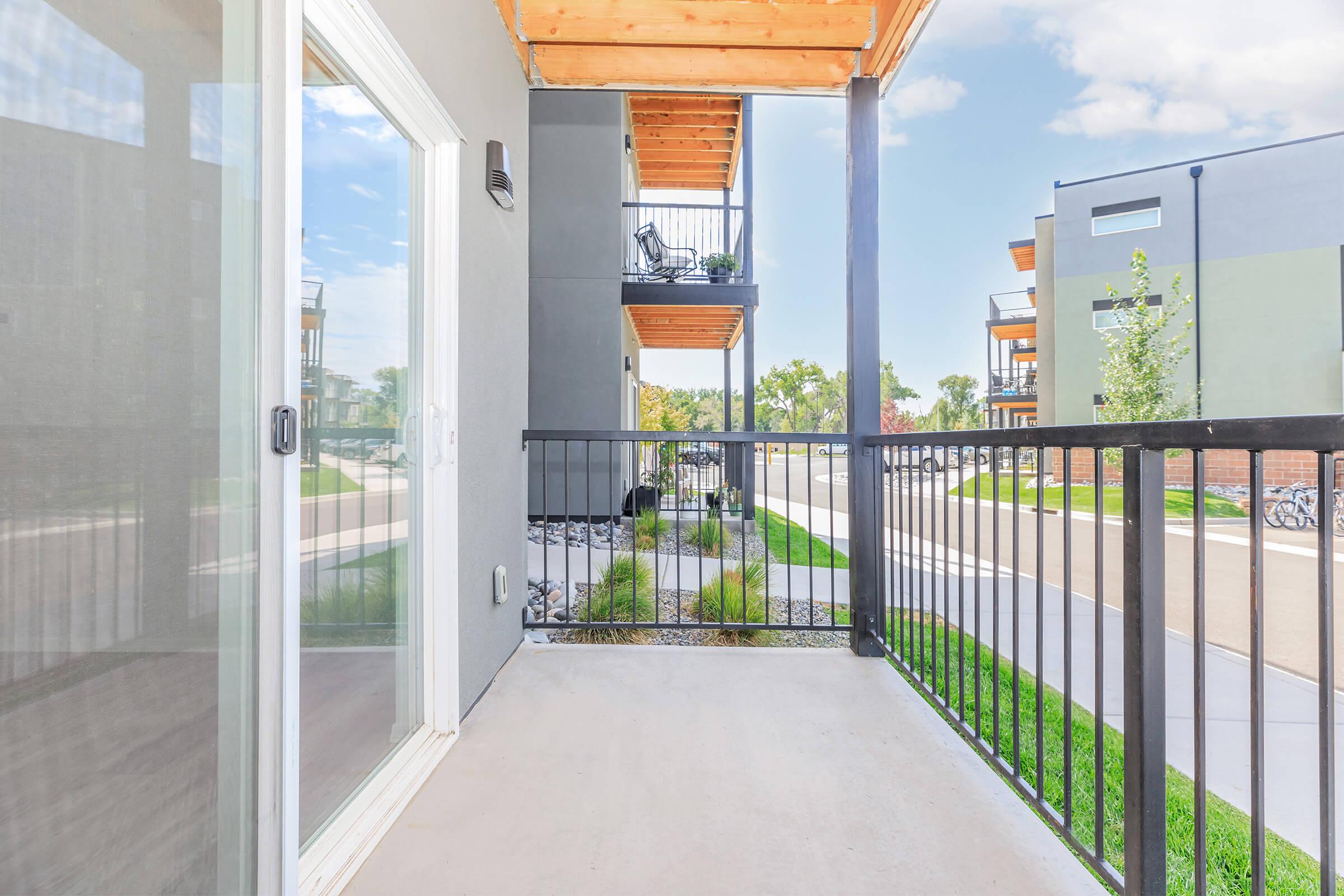 A view from a balcony of a modern apartment complex, featuring a concrete floor and black railings. In the foreground, there is a pathway, grass, and landscaping. Visible are other apartment buildings in the background under a clear blue sky.