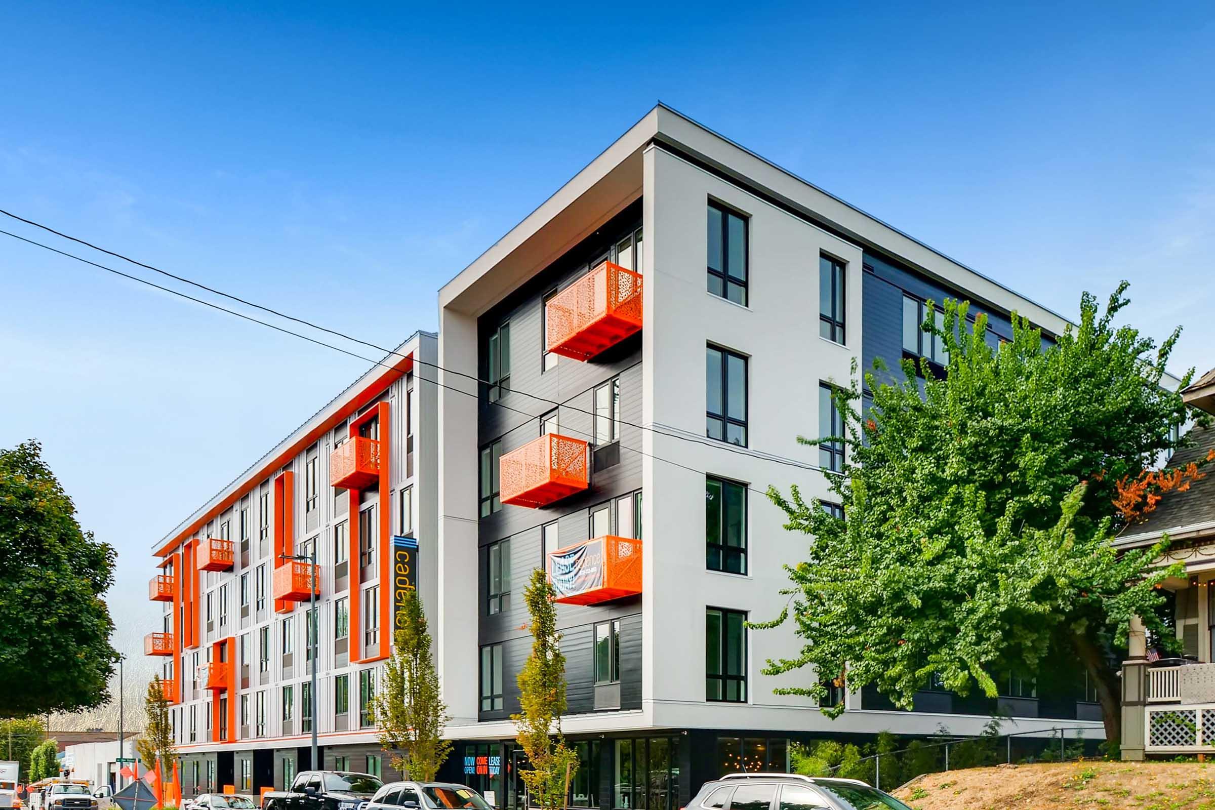 Modern multi-story apartment building featuring a mix of gray and orange exterior design. The structure has several balconies with orange railings, greenery around the base, and parked cars in the foreground. A clear blue sky serves as the backdrop.