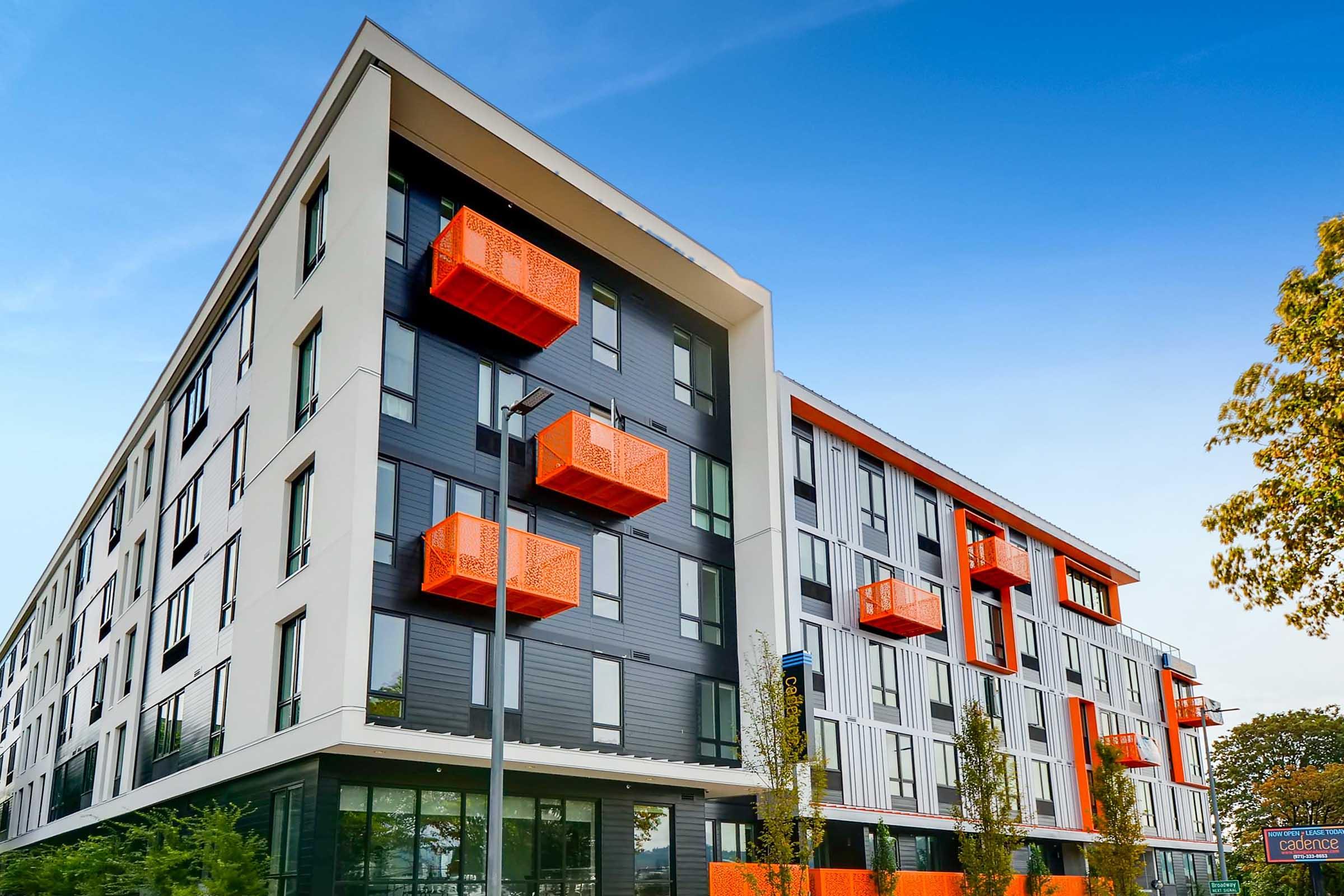 Modern multi-story building with a sleek design featuring a combination of dark and light-colored panels. Prominent orange balconies jut out from the facade, complemented by green landscaping and a clear blue sky in the background.