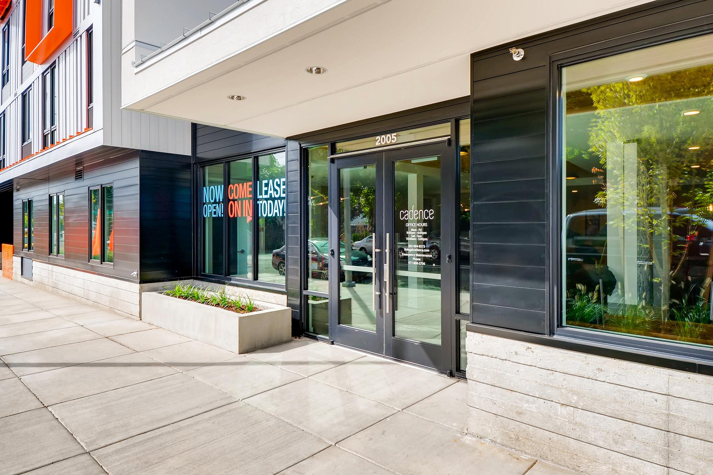 Exterior view of a modern commercial building with large glass doors and windows displaying colorful signs that read "NOW OPEN," "LEASE," and "COME IN TODAY." The entrance is well-lit and features clean lines, surrounded by landscaped greenery and a concrete pathway.