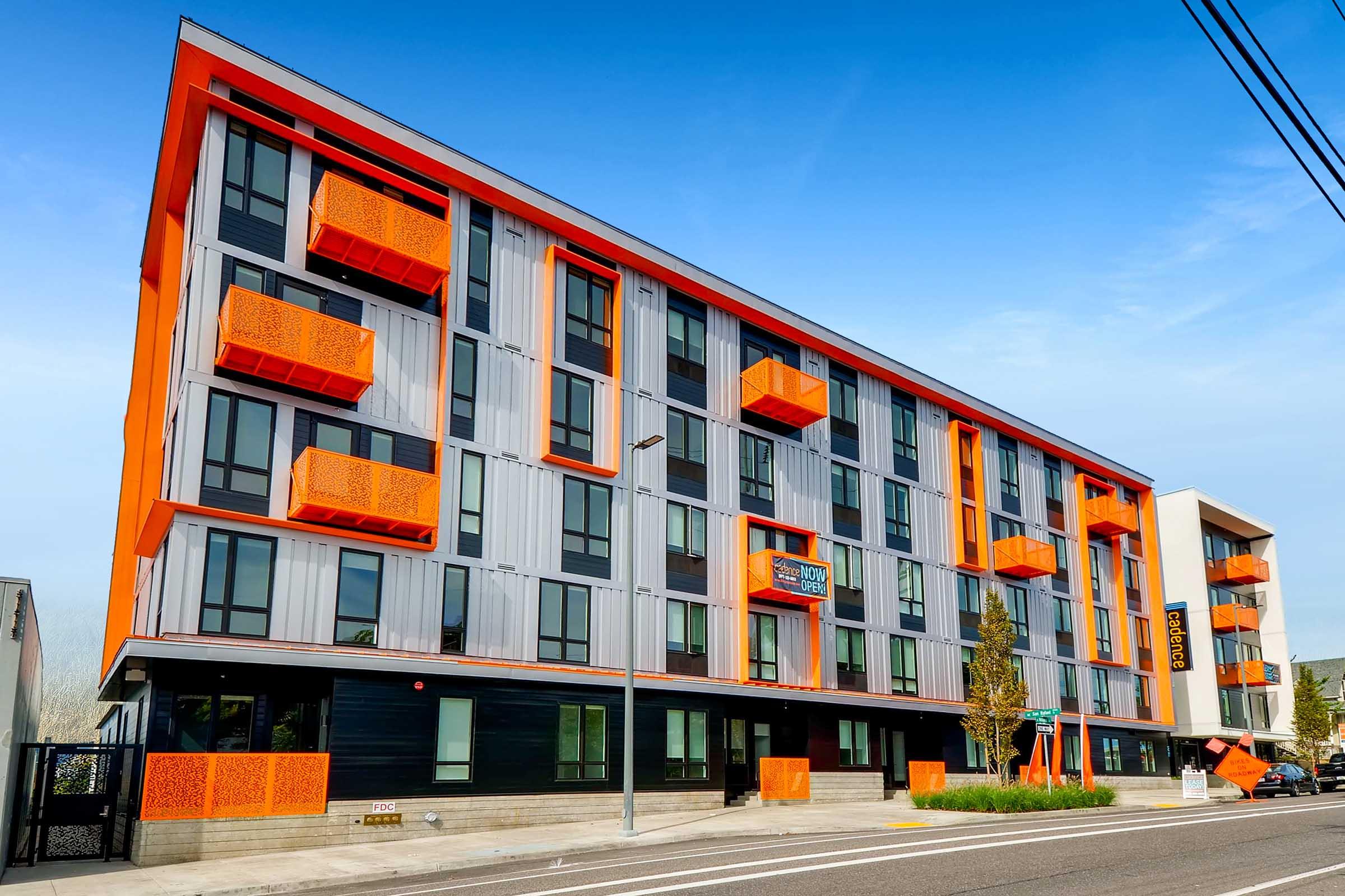 Modern building with a sleek design featuring a combination of gray and orange elements. The façade includes large orange balconies and decorative panels, with large windows. The structure has multiple floors and is situated at a street corner, under a clear blue sky.