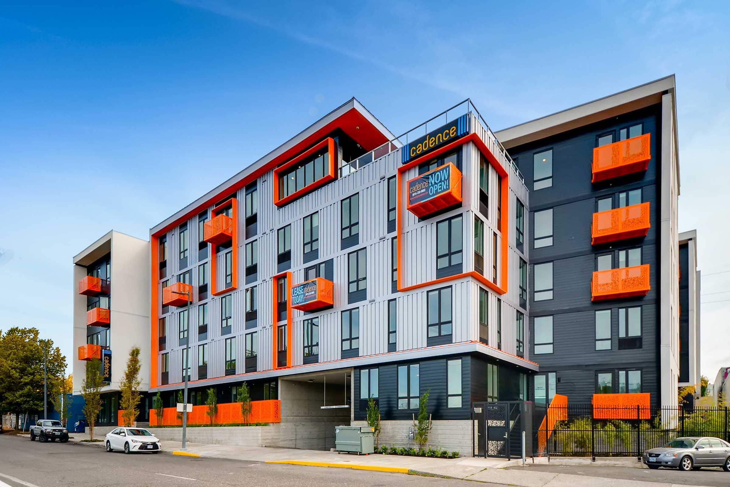 Modern multi-story apartment building with a sleek design featuring a combination of gray and vibrant orange accents. The building has large windows and balconies, with a spacious entrance area. Surrounding landscaping includes small trees and shrubs, enhancing the contemporary aesthetic. Clear blue sky above.