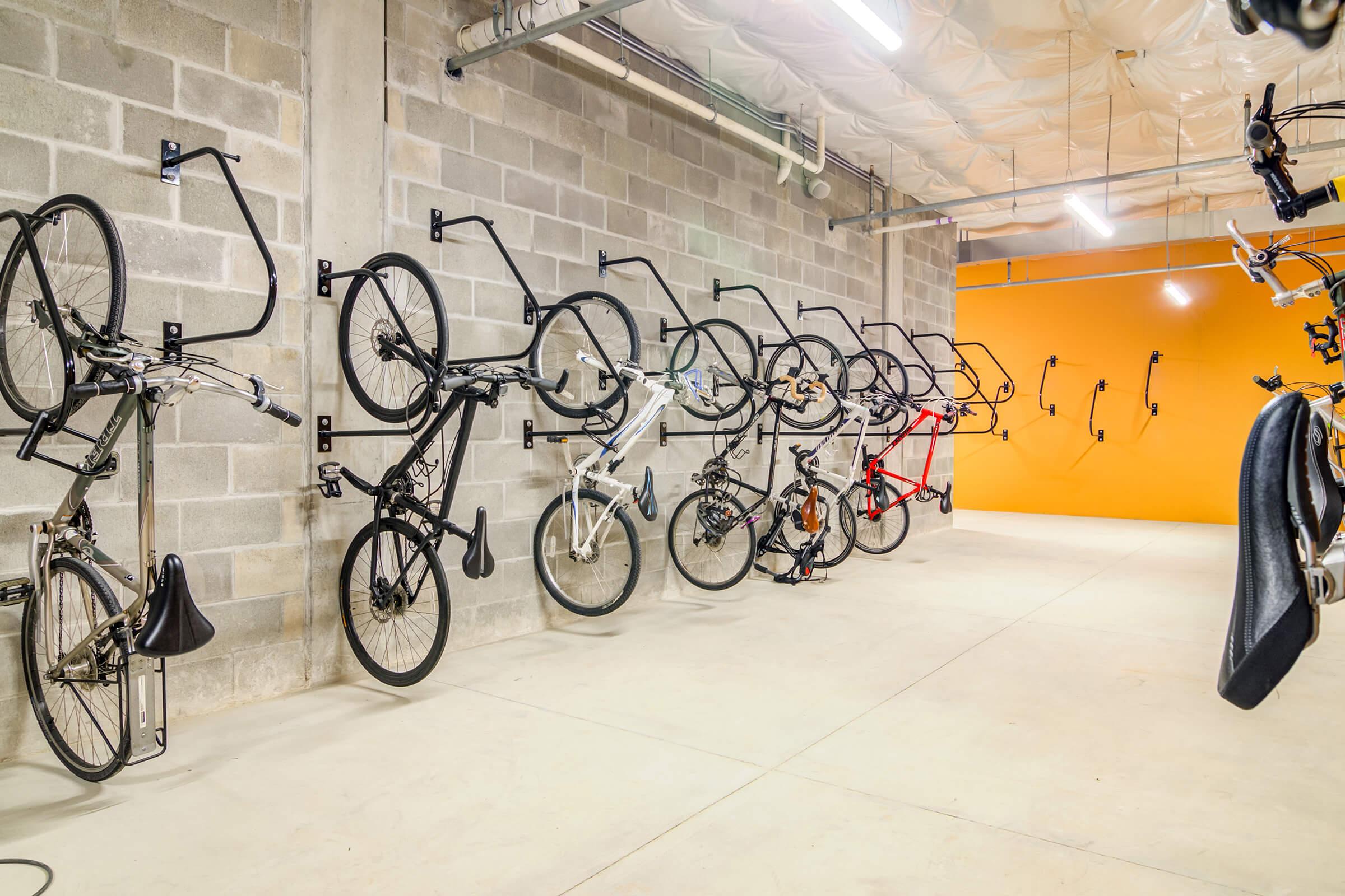 A modern bike storage area featuring several bicycles mounted on wall hooks, with a plain orange wall in the background. The floor is concrete, and the walls are made of grey cinder blocks, creating a clean and organized space for bike storage.