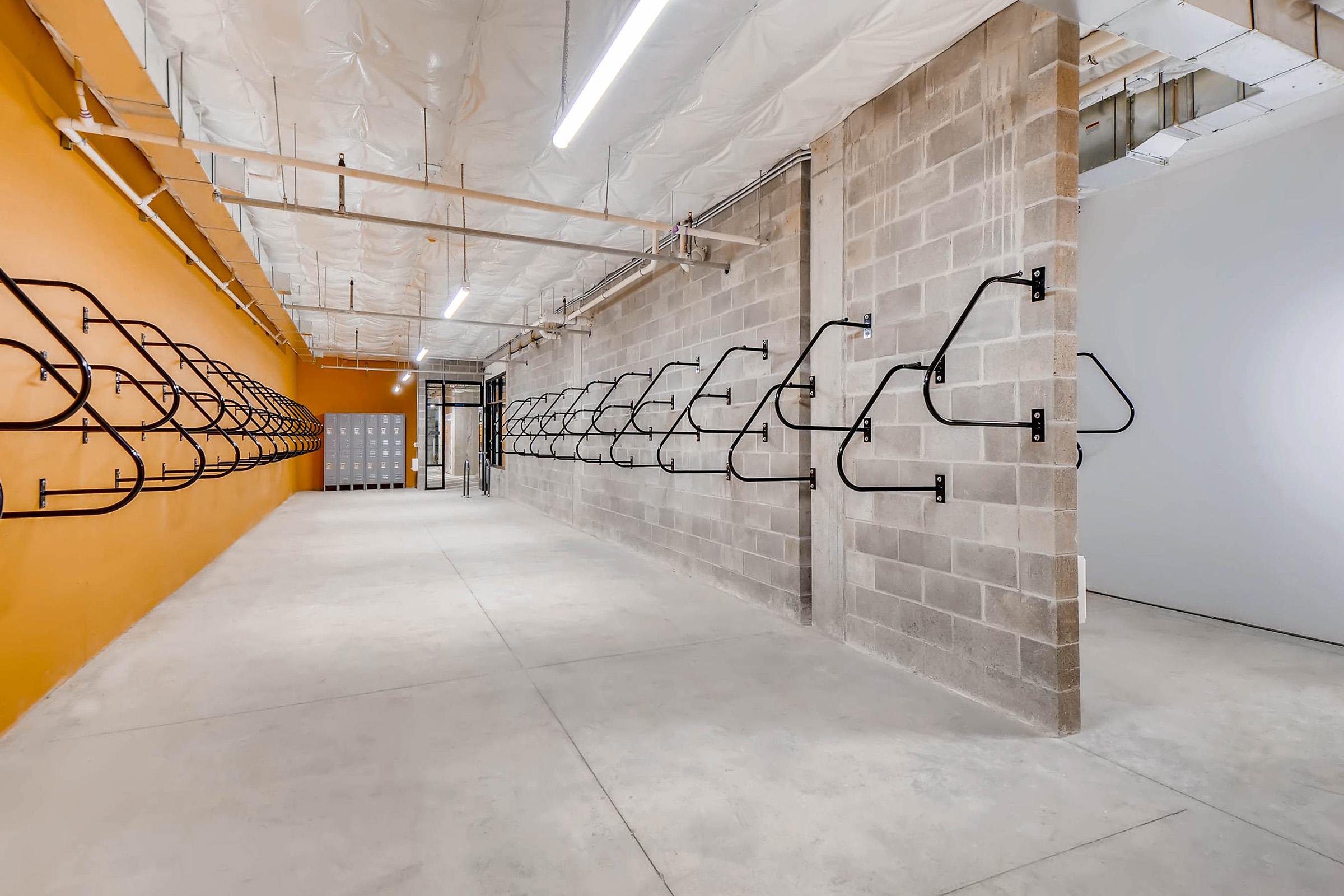 An interior space featuring exposed concrete walls with a bright orange accent wall. Metal bike racks are mounted along the walls. The area is well-lit with overhead lighting, and there is a row of lockers visible in the background on the left side. The floor is concrete and appears spacious.