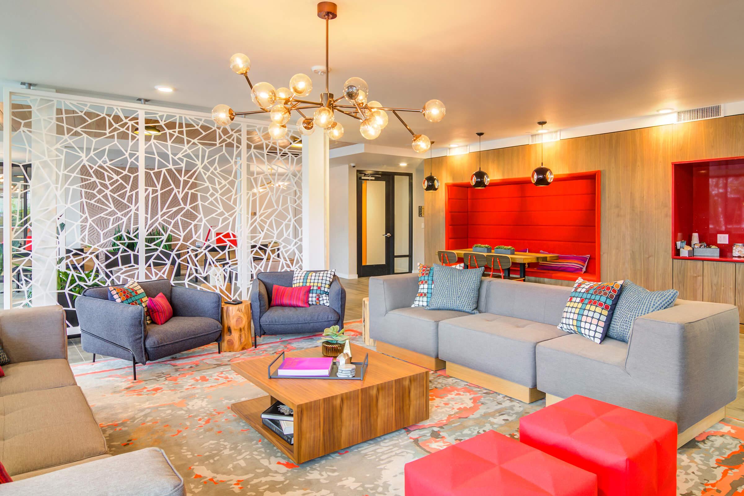 A modern living room featuring a mix of gray couches with colorful cushions, a wooden coffee table, and vibrant red ottomans. The space has decorative lighting, a patterned partition, and a bright area with a red accent wall. Soft lighting adds a cozy ambiance.