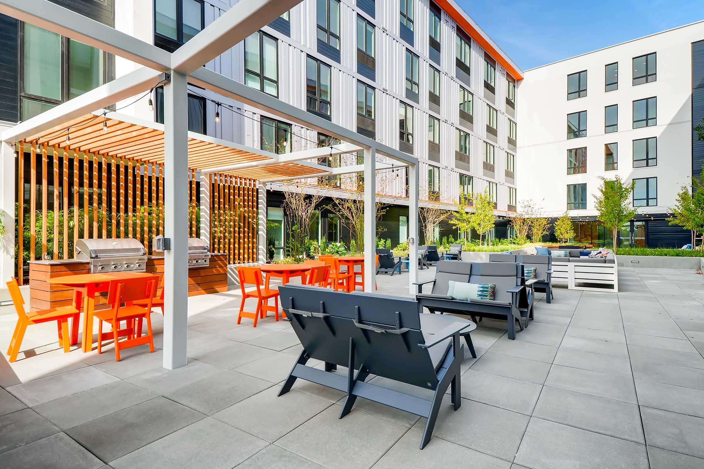 Outdoor communal area featuring modern furniture, including gray lounge chairs and orange dining tables. A grill station is visible, surrounded by neatly landscaped greenery. The backdrop consists of contemporary multi-story buildings with large windows and a clear blue sky.