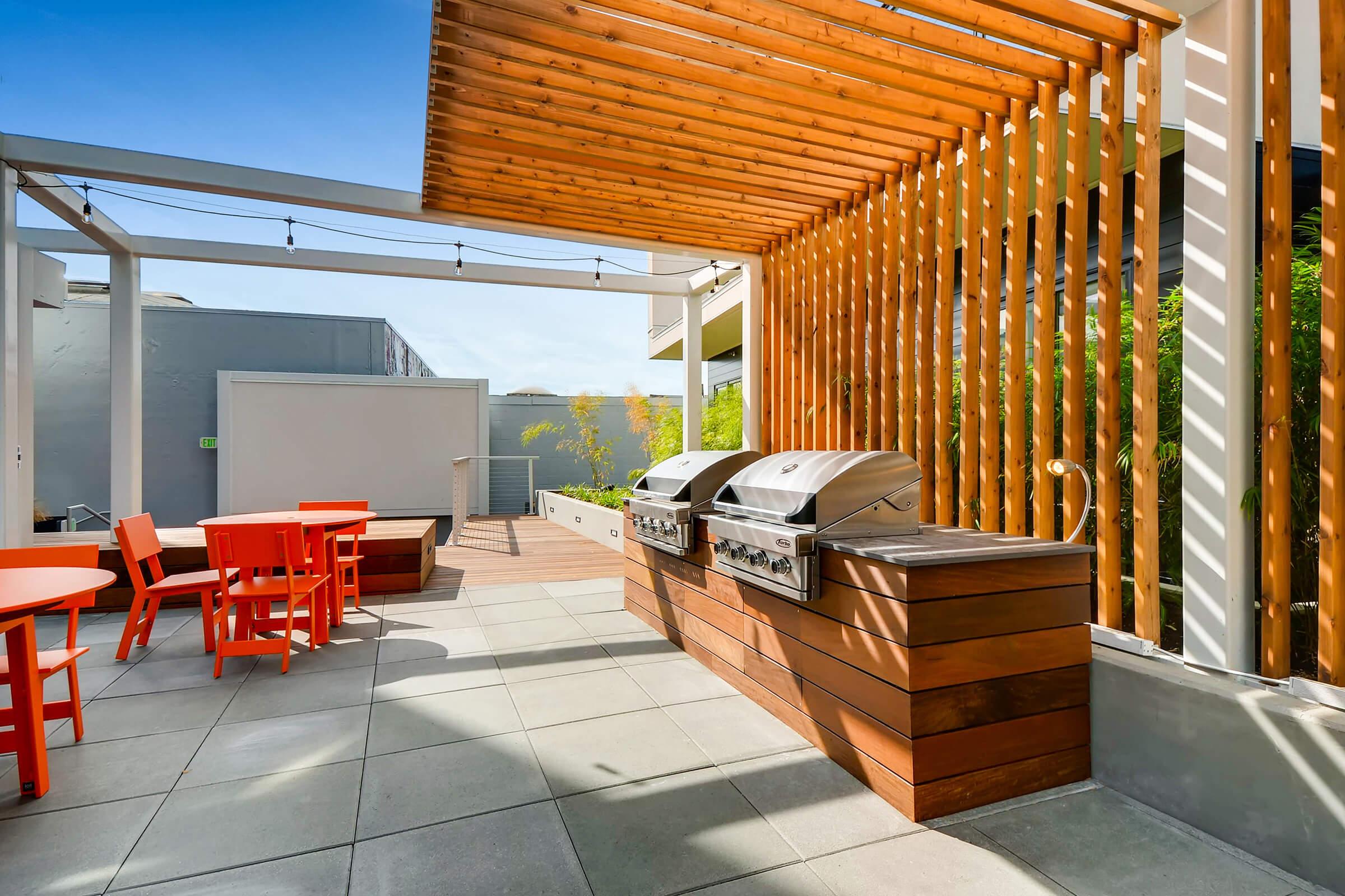 Outdoor grilling area featuring two stainless steel barbeque grills, wooden countertop, and vibrant orange furniture. The space is adorned with a modern wooden slatted structure and lush greenery, providing a stylish and inviting atmosphere for gatherings. Clear blue sky overhead enhances the outdoor setting.