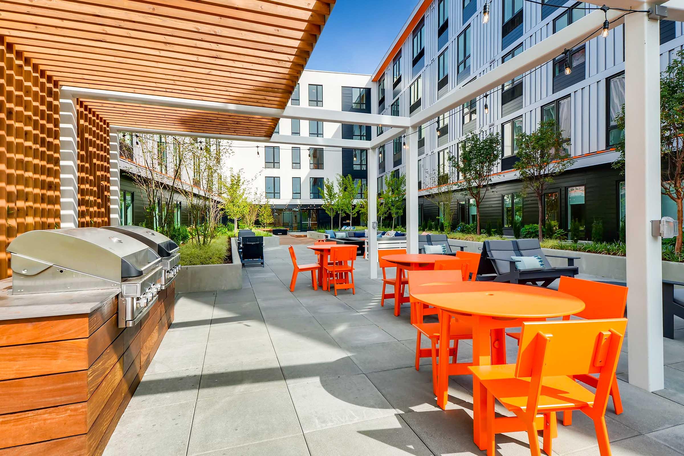 Outdoor communal area featuring vibrant orange tables and chairs, a stainless steel grill, and landscaped greenery. The backdrop includes a modern building with large windows and a blue sky, creating a welcoming atmosphere for social gatherings.