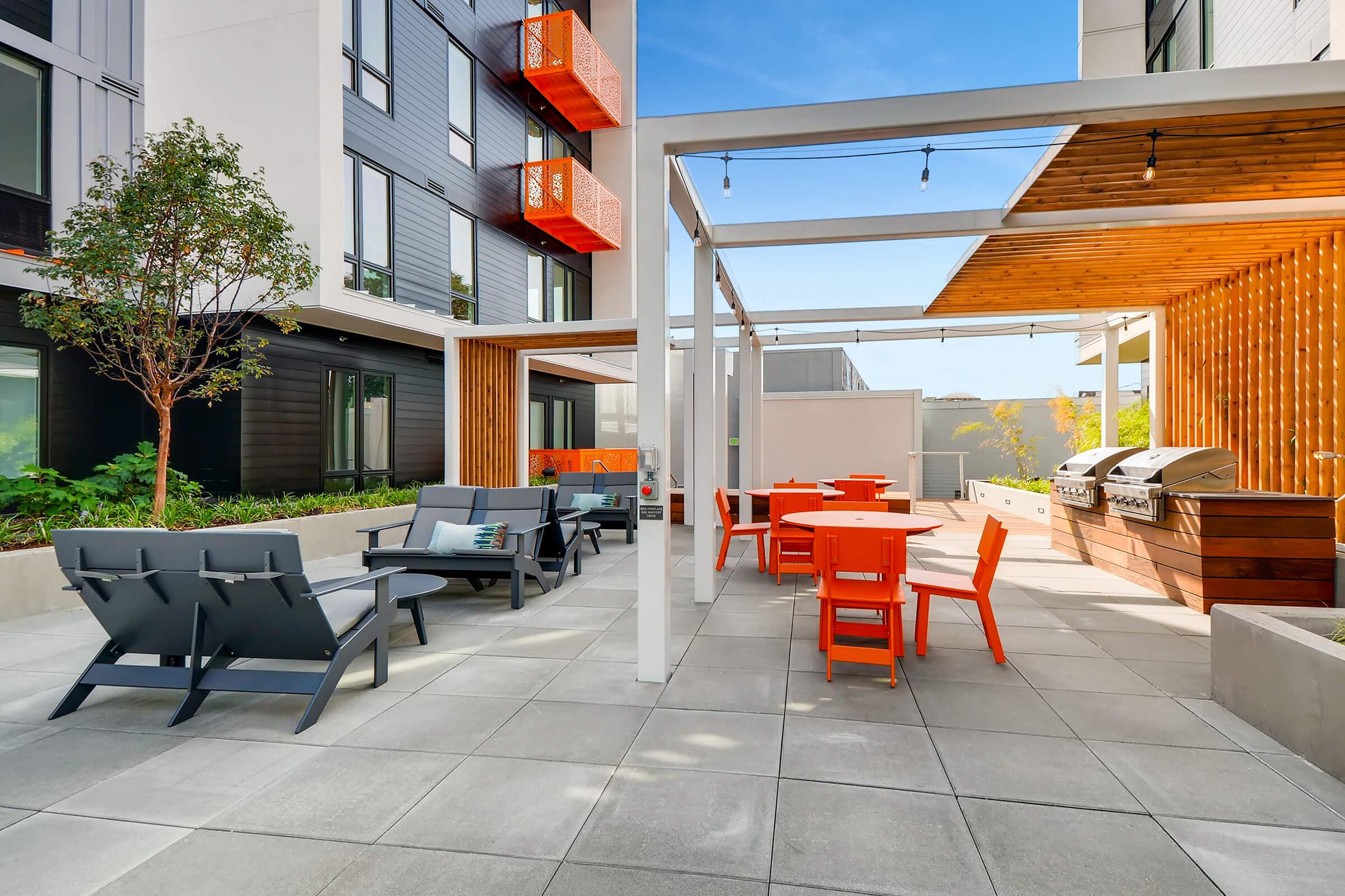 A modern outdoor patio area featuring comfortable black lounge chairs, circular orange tables, and a barbecue grill. The space is well-lit with string lights and surrounded by greenery, offering a welcoming atmosphere for relaxation and social gatherings.