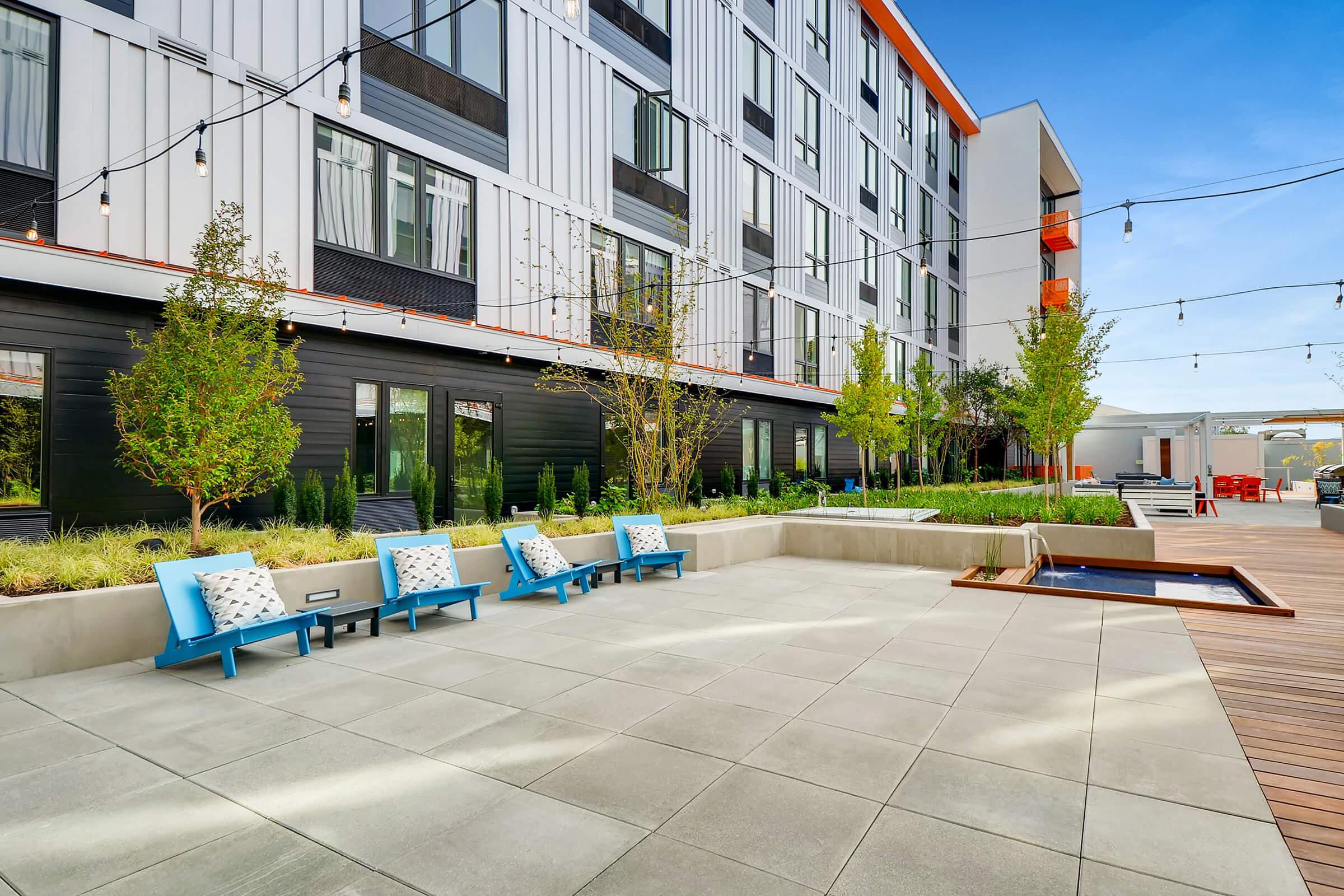 A modern outdoor patio with several blue lounge chairs, surrounded by greenery and decorative plants. In the background, a contemporary building with large windows and orange accents is visible. String lights hang above, adding ambiance to the outdoor space.