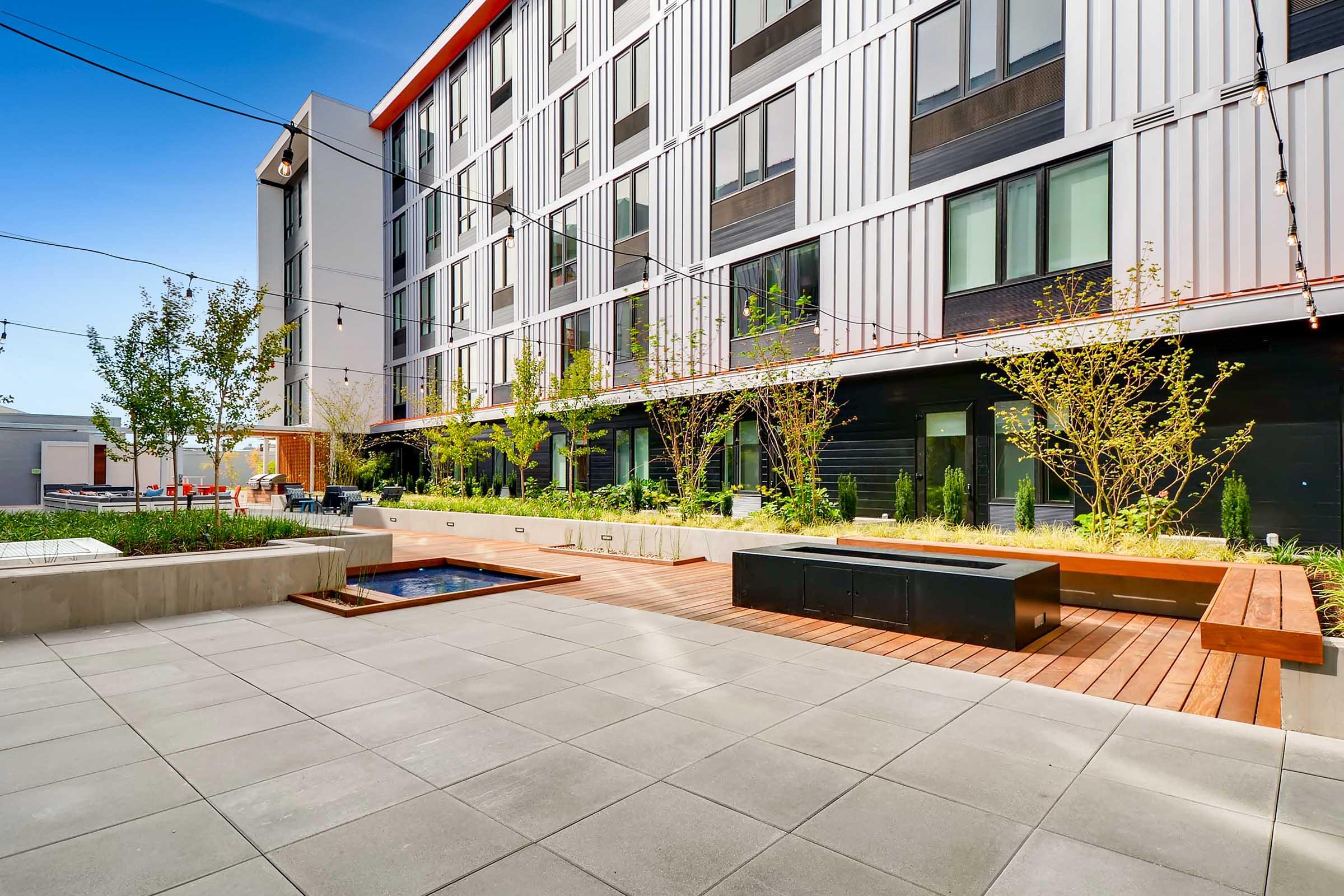 A modern courtyard featuring a mix of concrete and wooden elements. There are landscaped areas with small trees and shrubs, a shallow water feature, and seating areas made of wood. The backdrop consists of a stylish multi-story building with large windows and a red roof. String lights add ambiance to the space.