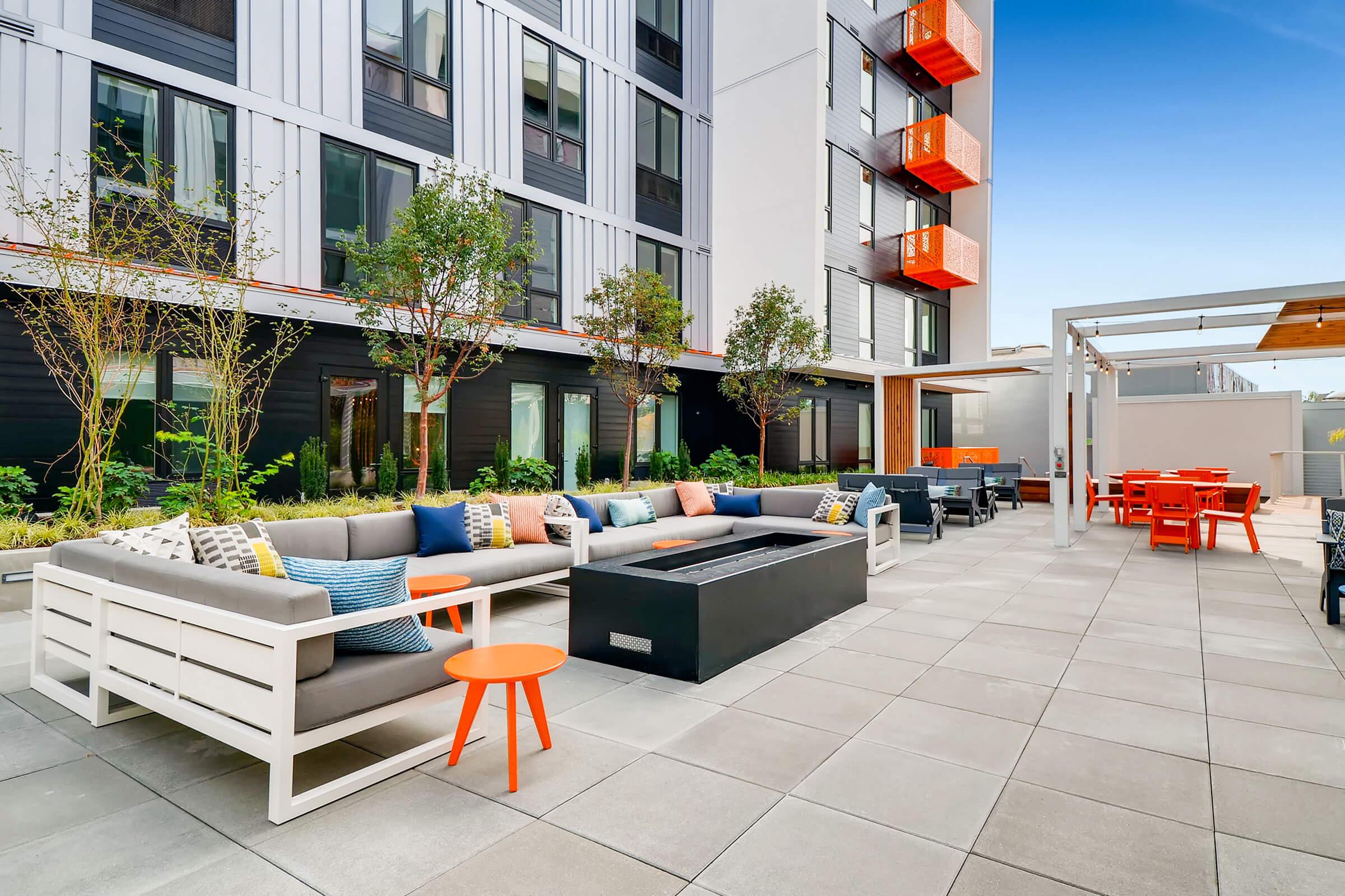 Modern outdoor seating area with a large sectional sofa adorned with colorful cushions, an orange accent table, and a fire pit. The space features contemporary architecture with green trees and shrubs, along with a dining area and bright orange balconies in the background, set against a clear blue sky.