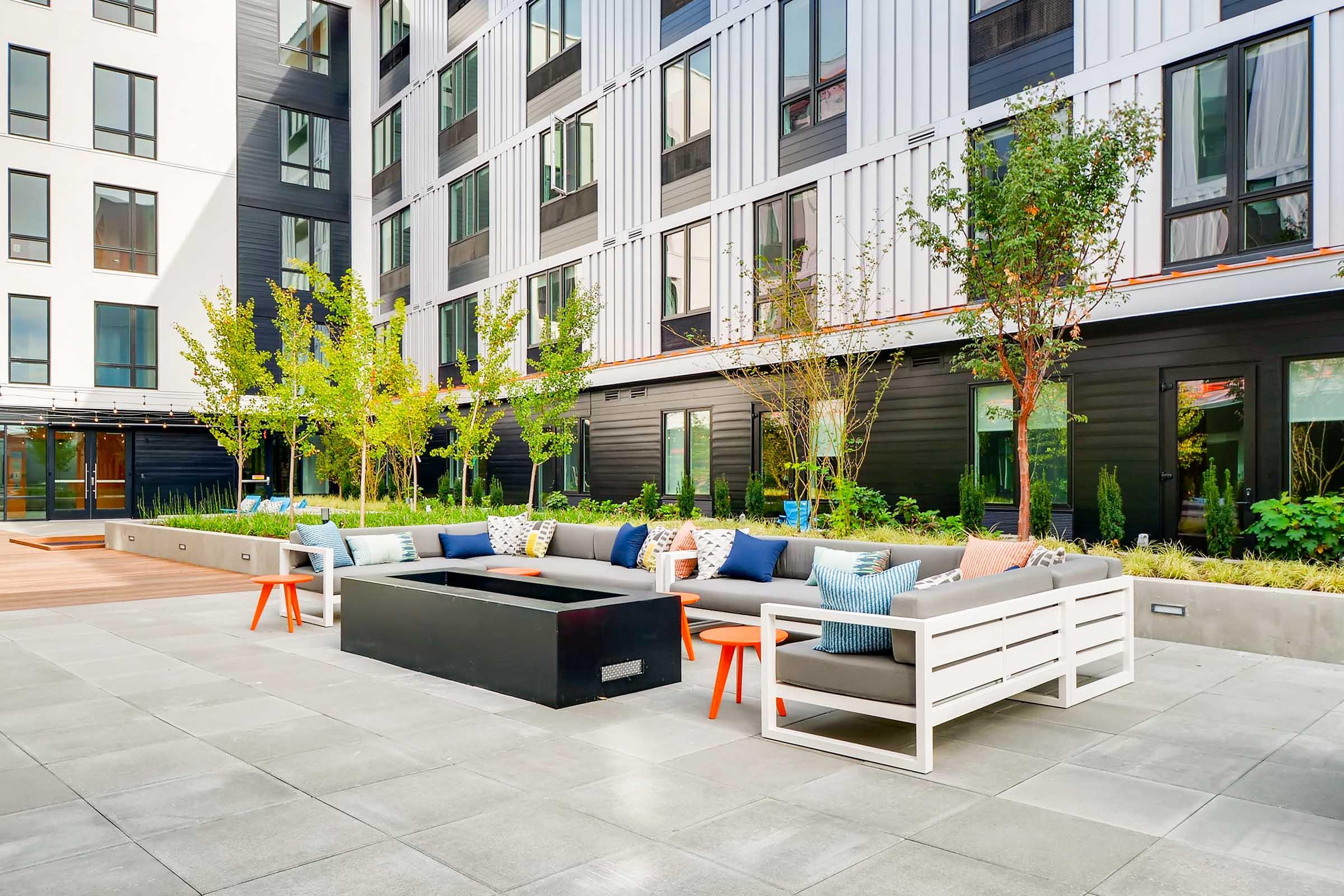 A modern outdoor courtyard featuring a small water feature surrounded by wooden decking. Lush greenery and plants line the space, with contemporary apartment buildings in the background. Strung overhead lights add a warm ambiance to the inviting area, ideal for relaxation or gatherings.