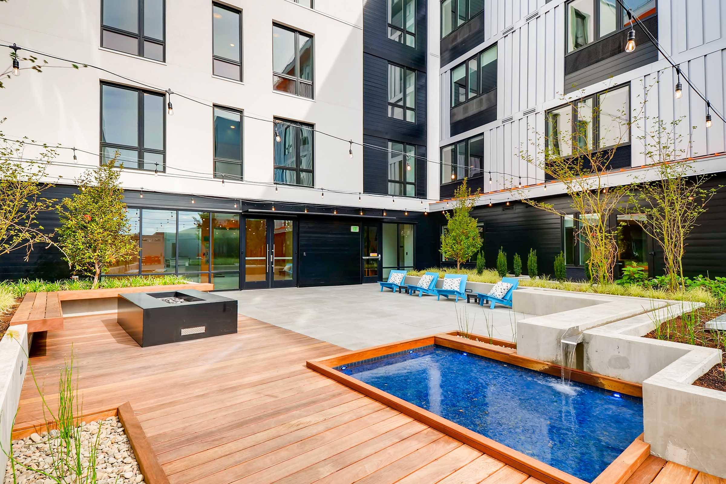 A modern outdoor courtyard featuring a wooden deck, a small blue pool, and a fire pit. Surrounding the area are potted plants and lounge chairs. The building has large windows, with a sleek contemporary design, and string lights overhead create a cozy atmosphere.