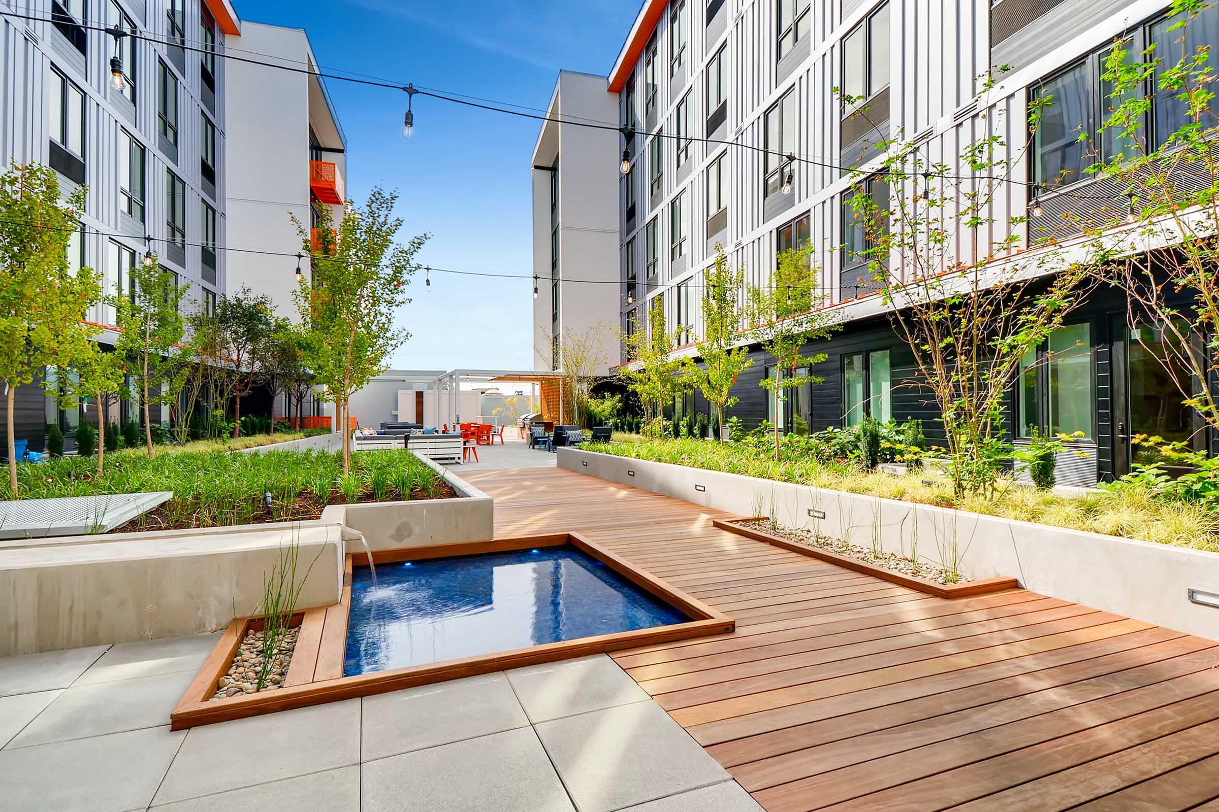 A modern outdoor lounge area featuring a large sectional sofa with colorful cushions and orange accent tables. There are several trees and plants surrounding the space, alongside contemporary architecture in the background. The area is well-lit and invites relaxation and social gatherings.