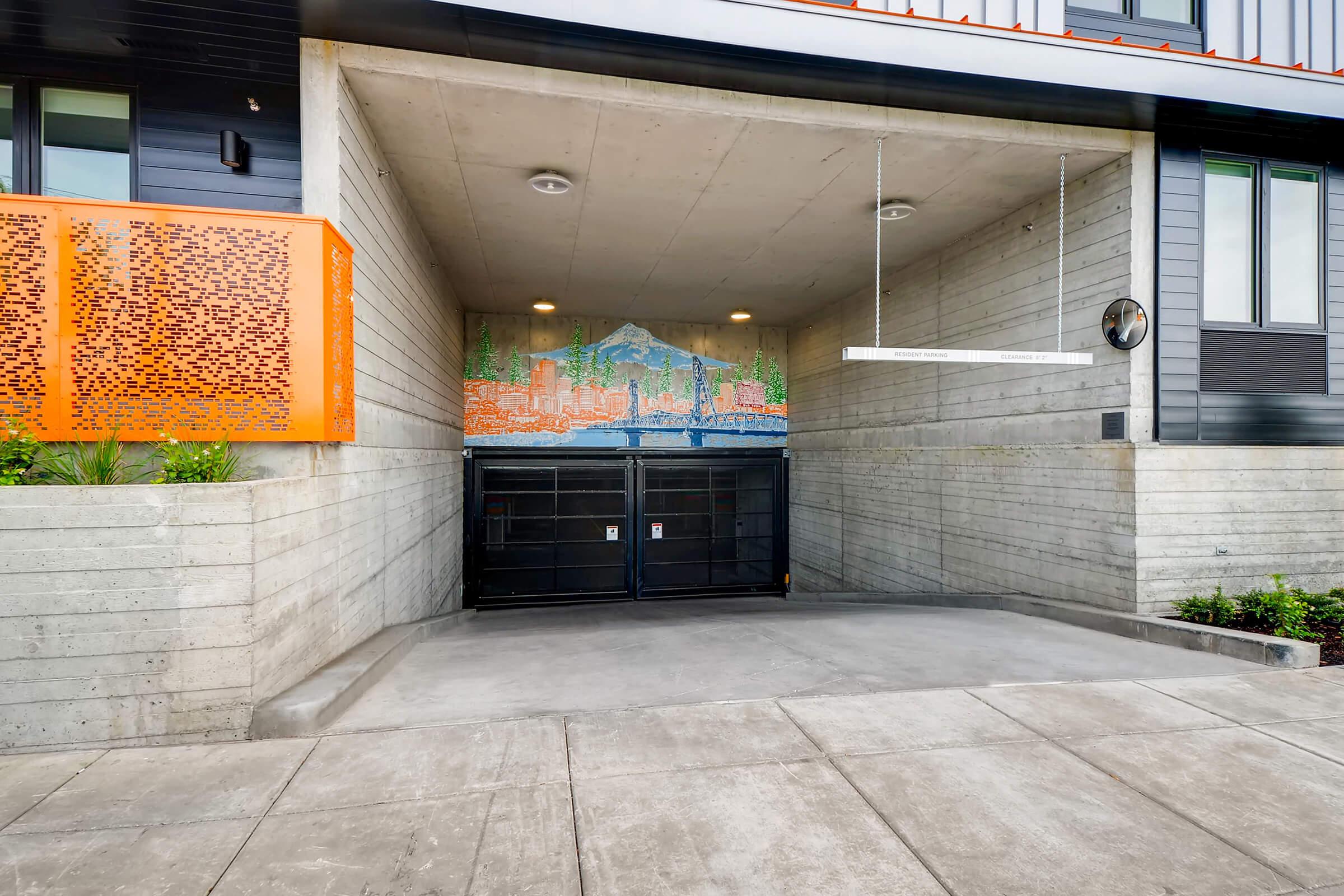 A concrete garage entrance with a mural of a city skyline in vibrant colors on the back wall. The entrance features a black gate and modern architectural elements, surrounded by neatly landscaped greenery. Light fixtures are installed on the ceiling for illumination.