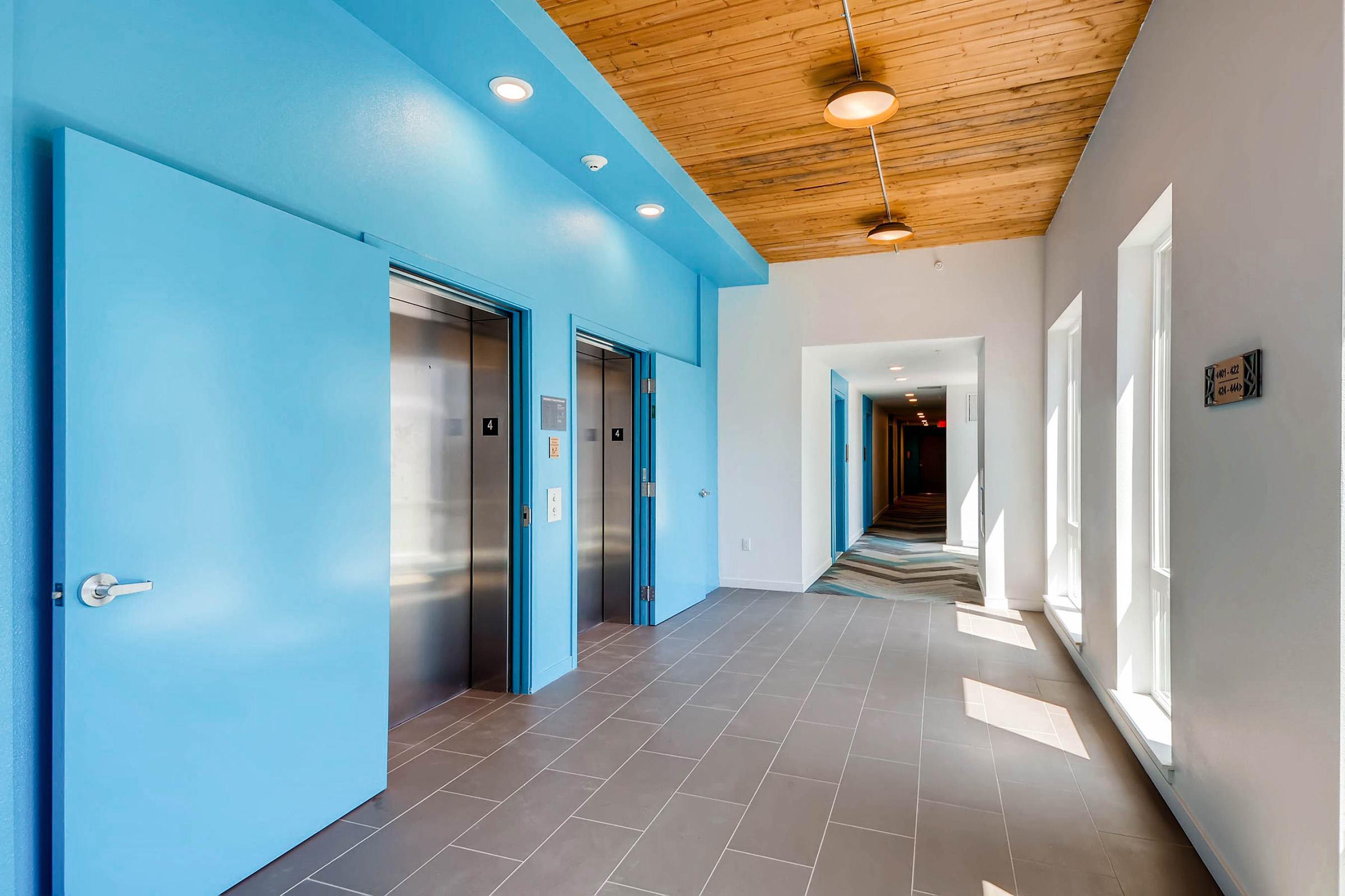 Brightly lit hallway featuring two blue elevator doors on one side and large windows. The ceiling has wooden panels, and there are modern light fixtures. The floor is tiled in a neutral color, and a patterned carpet is visible further down the corridor.