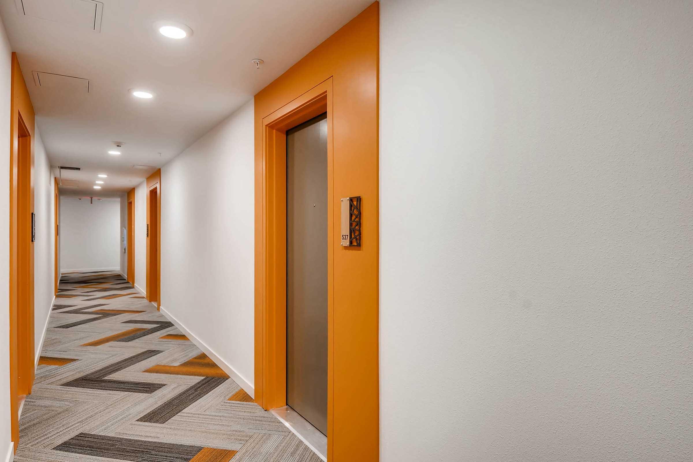 A well-lit hallway with orange accents and a patterned carpet. Two elevators are visible along the wall, with doors painted orange. The walls are a light color, creating a modern and welcoming atmosphere.
