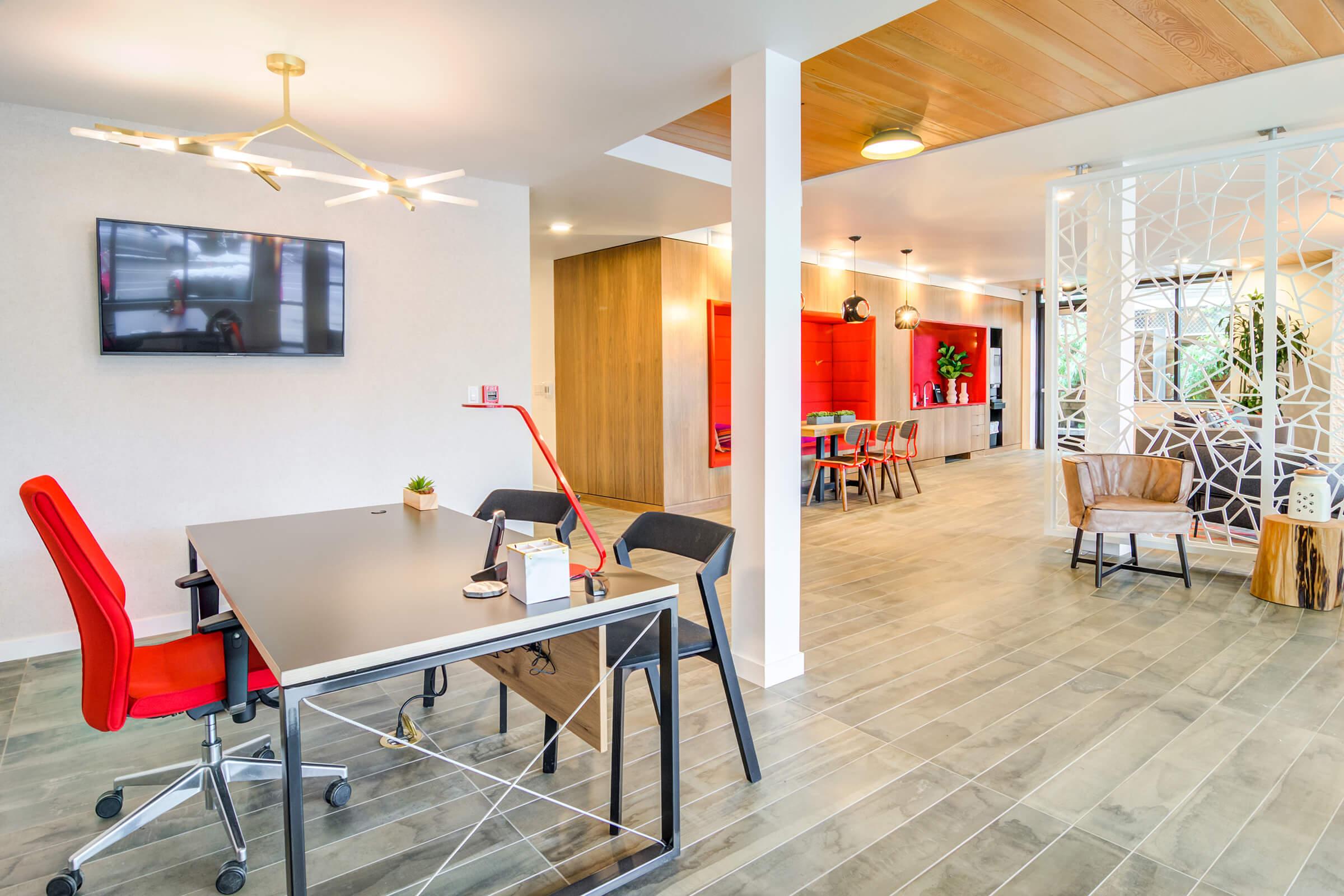 Modern office interior featuring a sleek desk with a red chair, a wall-mounted television, and stylish decor. The space has large windows, wooden accents, and a contemporary design, with plants and colorful artwork enhancing the ambiance. A bright, inviting atmosphere promotes collaboration and productivity.