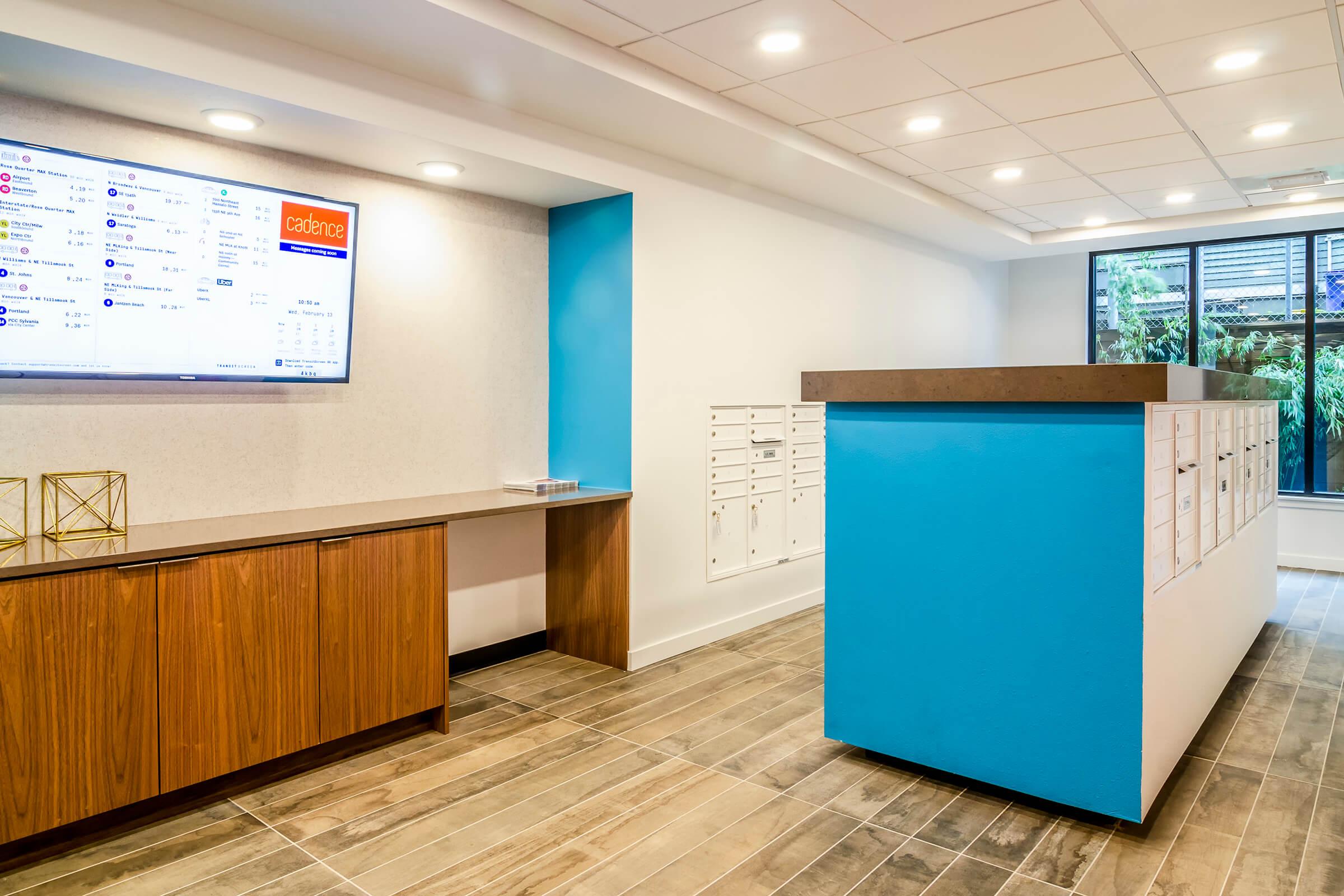 Modern lobby area featuring a sleek reception desk with a large wall-mounted screen displaying information. To the right, there are individual mailboxes organized neatly. The color scheme includes light wood tones and bright blue accents, complemented by recessed lighting and large windows allowing natural light.