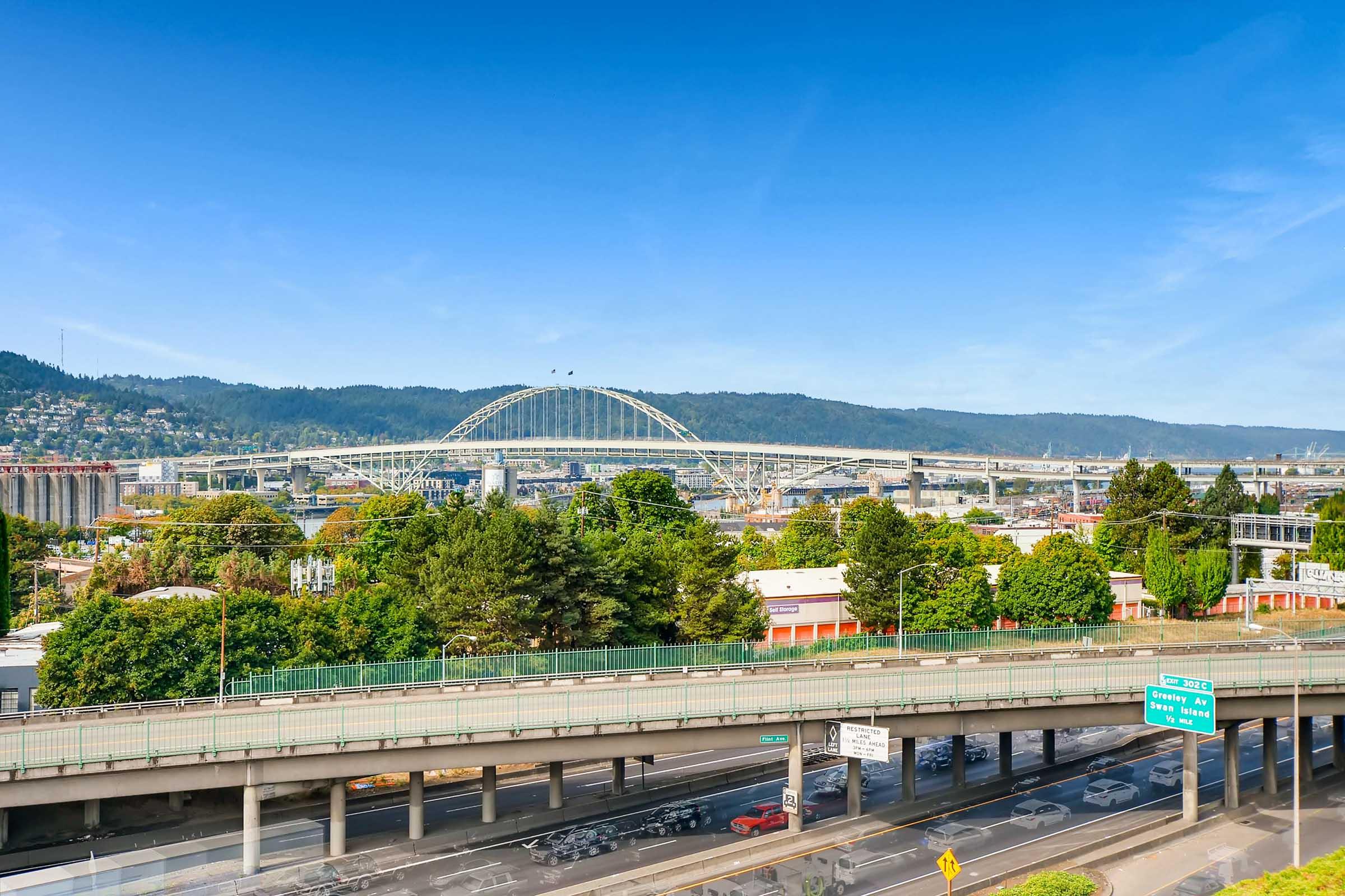 A scenic view of a cityscape featuring a bridge arching over a river, surrounded by lush greenery and hills. Below, a highway with vehicles traversing, flanked by trees and urban buildings, under a clear blue sky. The image captures the blend of nature and infrastructure.